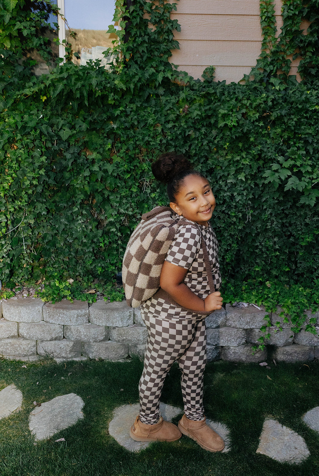 Side and back view of a child wearing brown check pajamas and a matching quillow backpack.