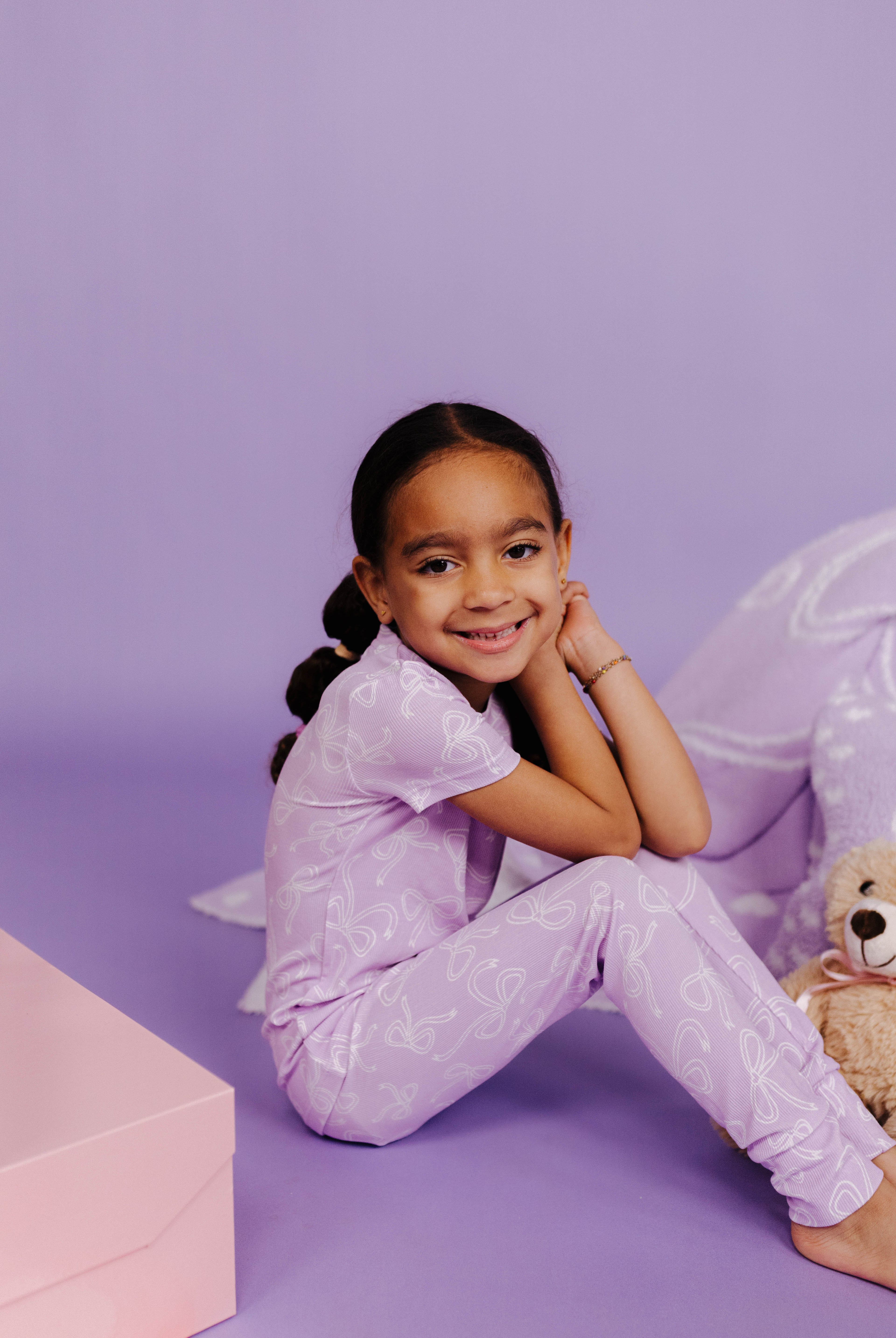 Young girl in a purple outfit sitting on a purple floor with a teddy bear.