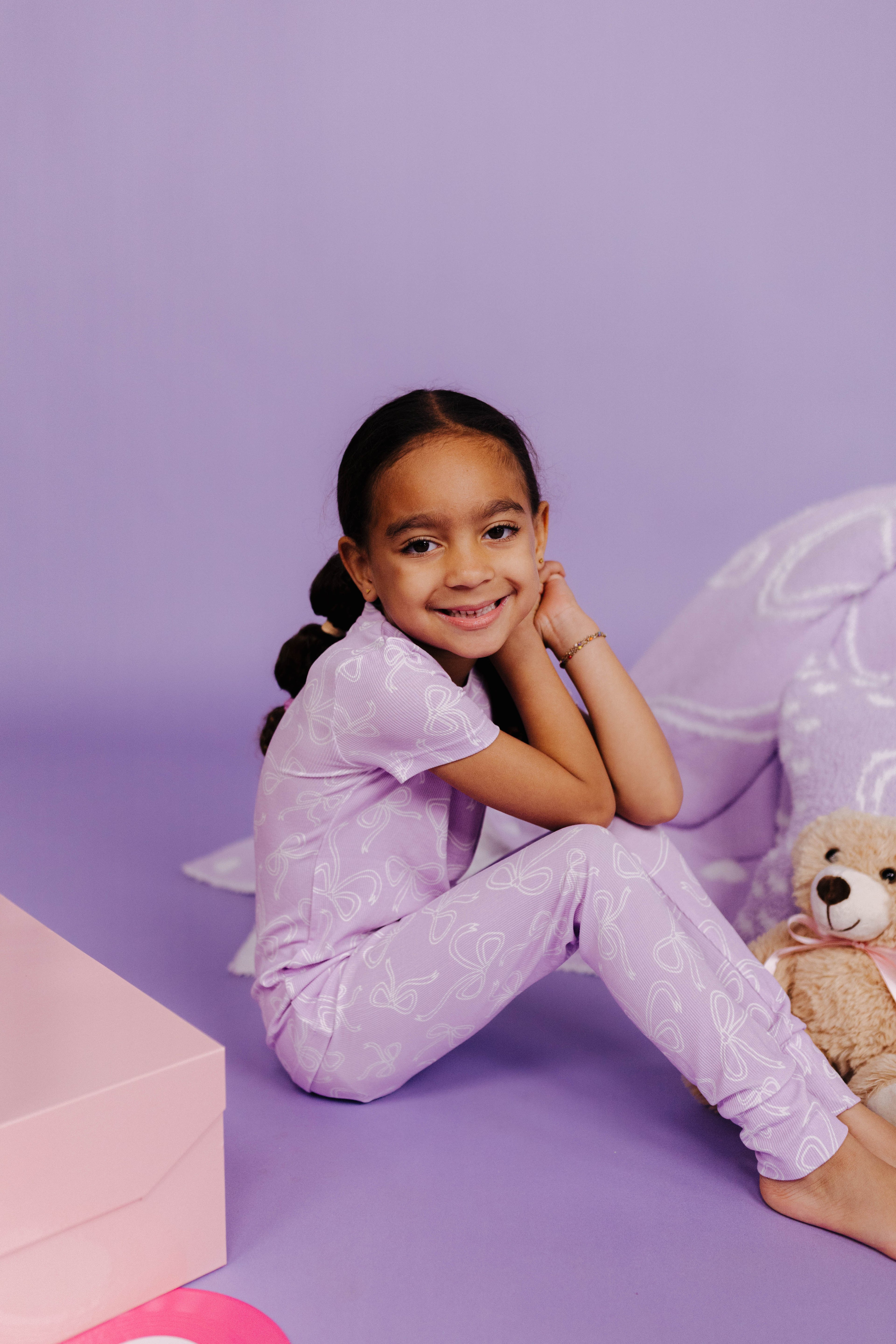 Young girl in a purple outfit sitting on a purple floor with a teddy bear.