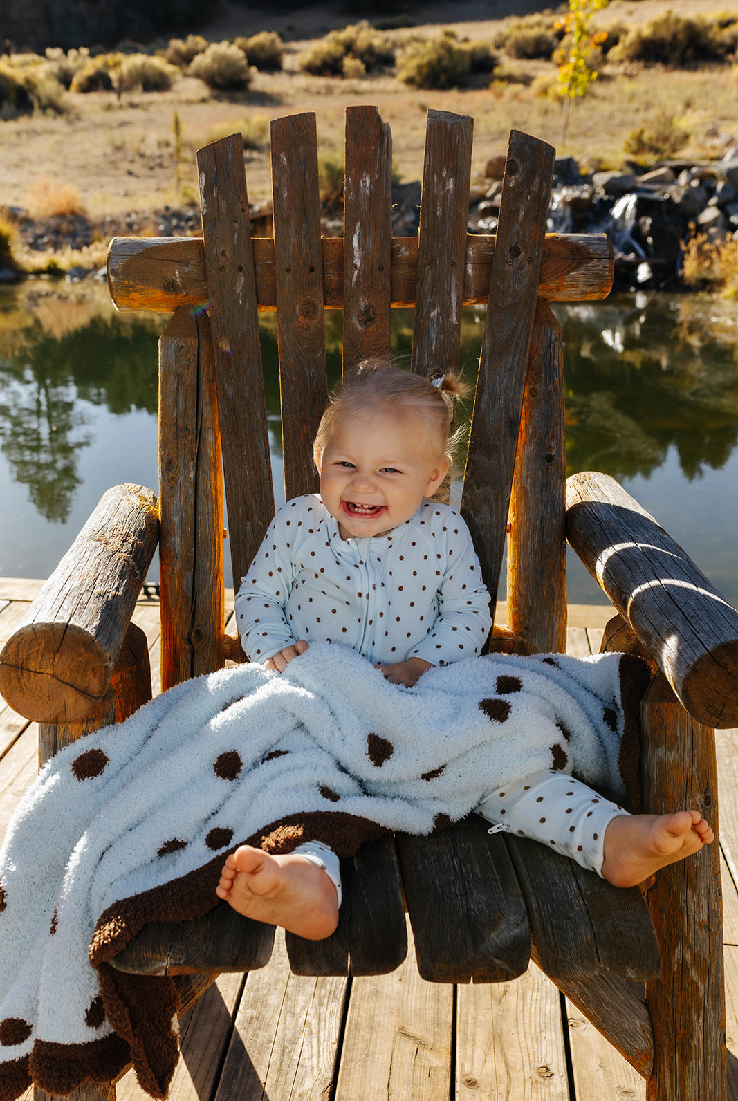 Baby seated in a wooden chair with a blue and brown polka dot kid blanket across their lap.