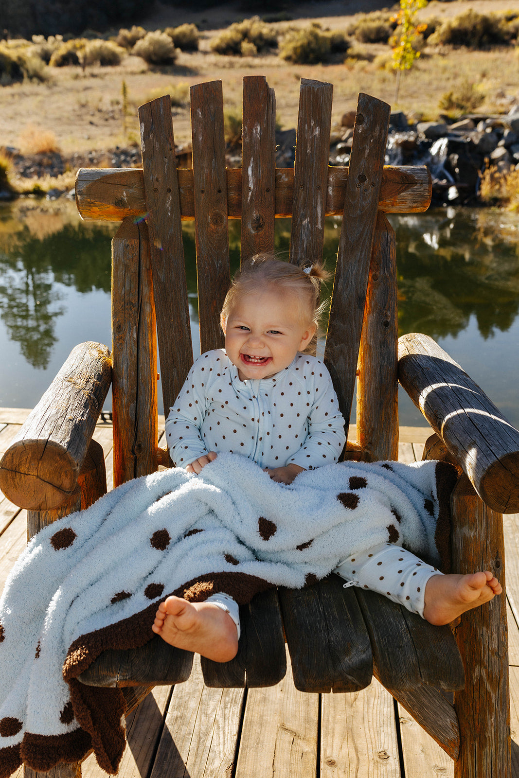 Baby seated in a wooden chair with a blue and brown polka dot kid blanket across their lap.