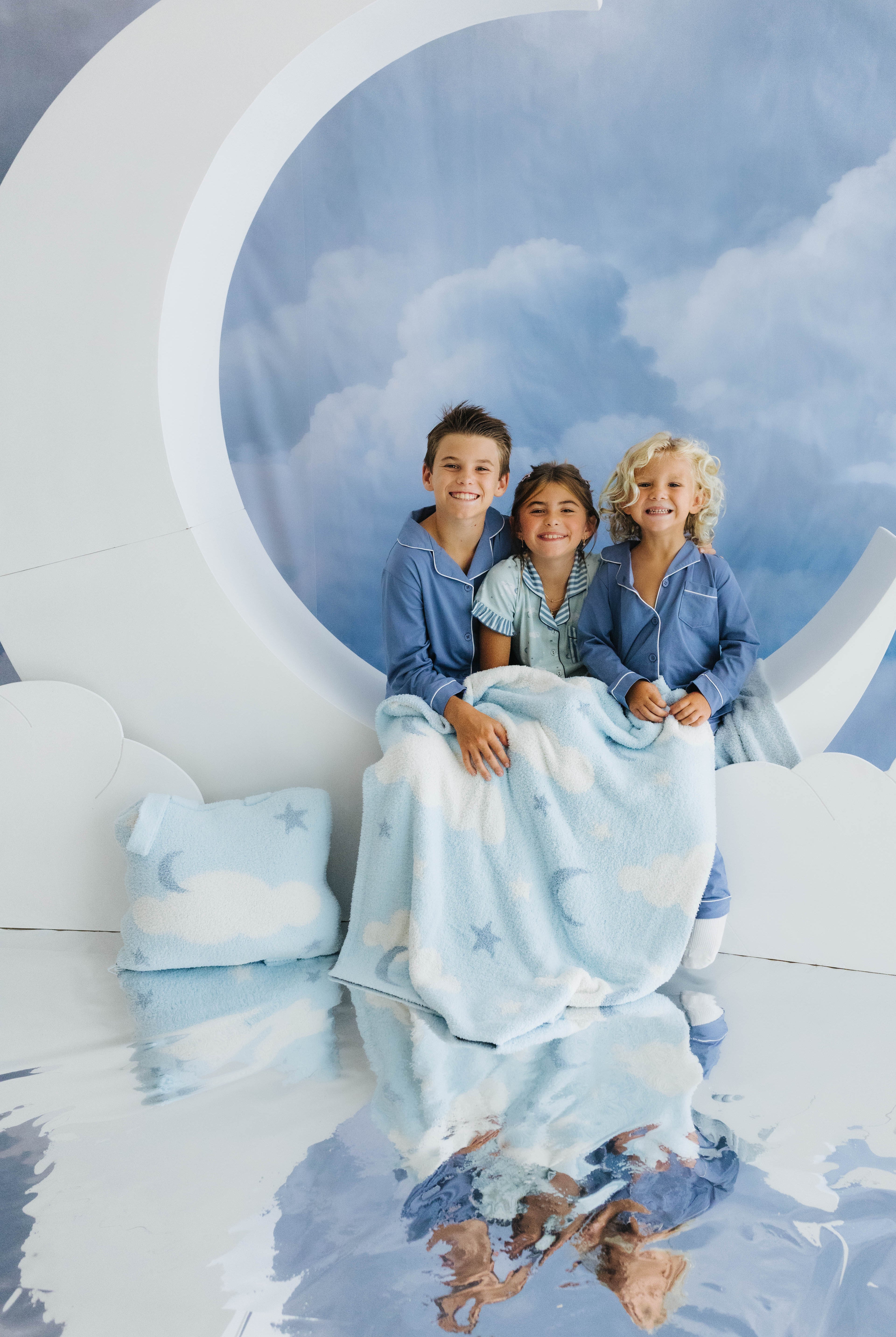 three kids smiling sitting on moon decoration with the baby blanket on their laps and cloud backpack/wristlet quillow on the side