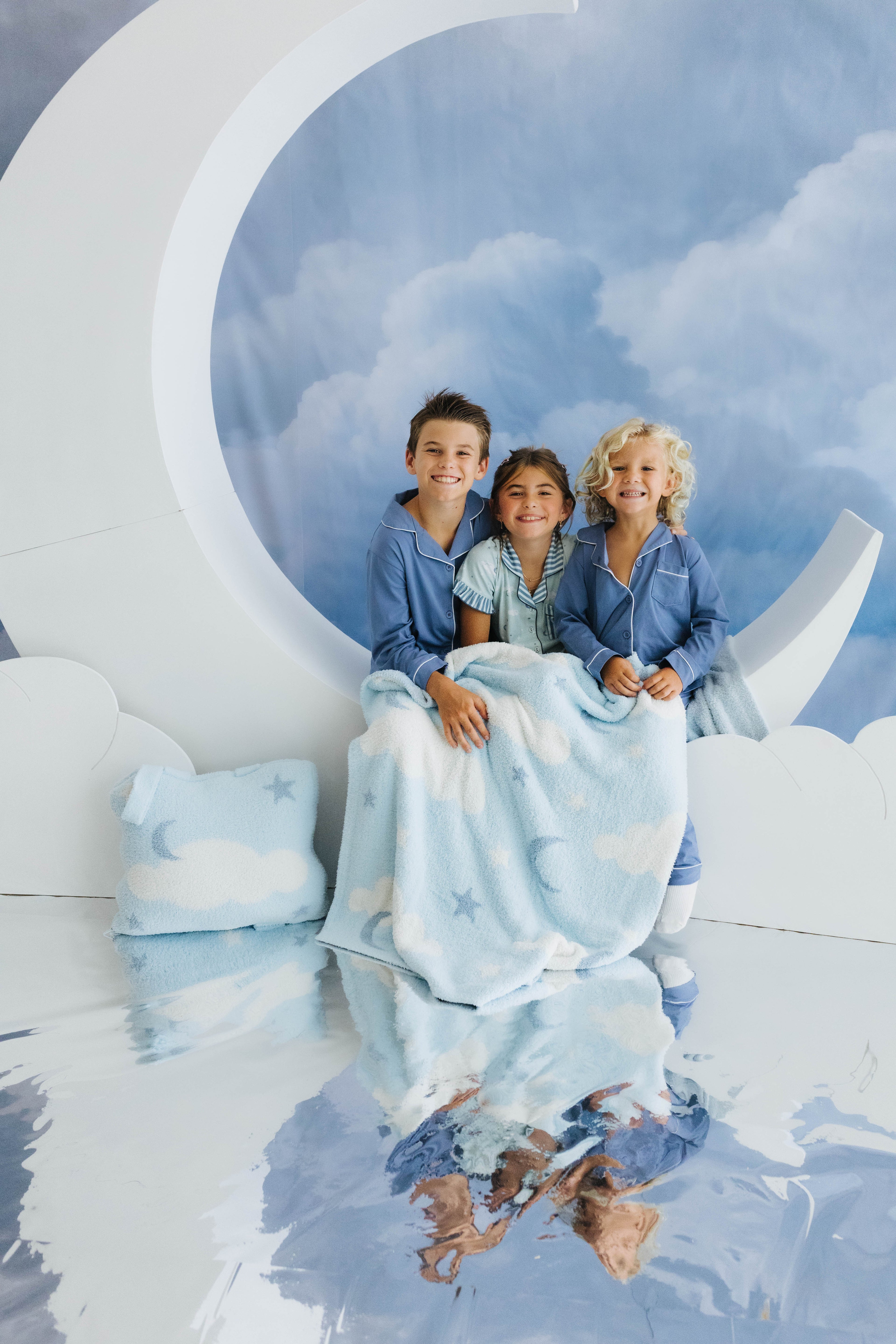 three kids smiling sitting on moon decoration with the baby blanket on their laps and cloud backpack/wristlet quillow on the side