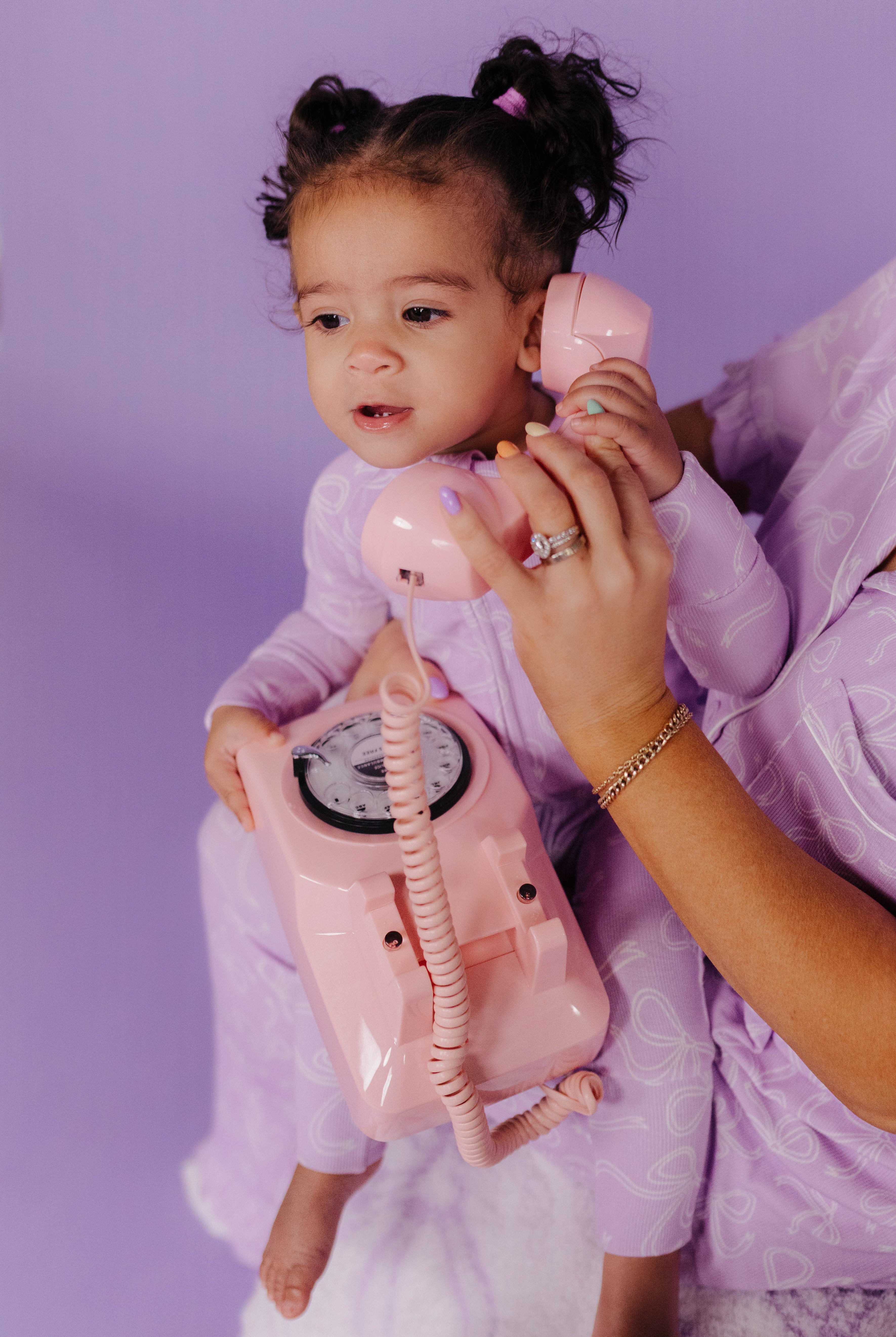 Child holding a pink vintage-style telephone with a person's hand, all against a purple background