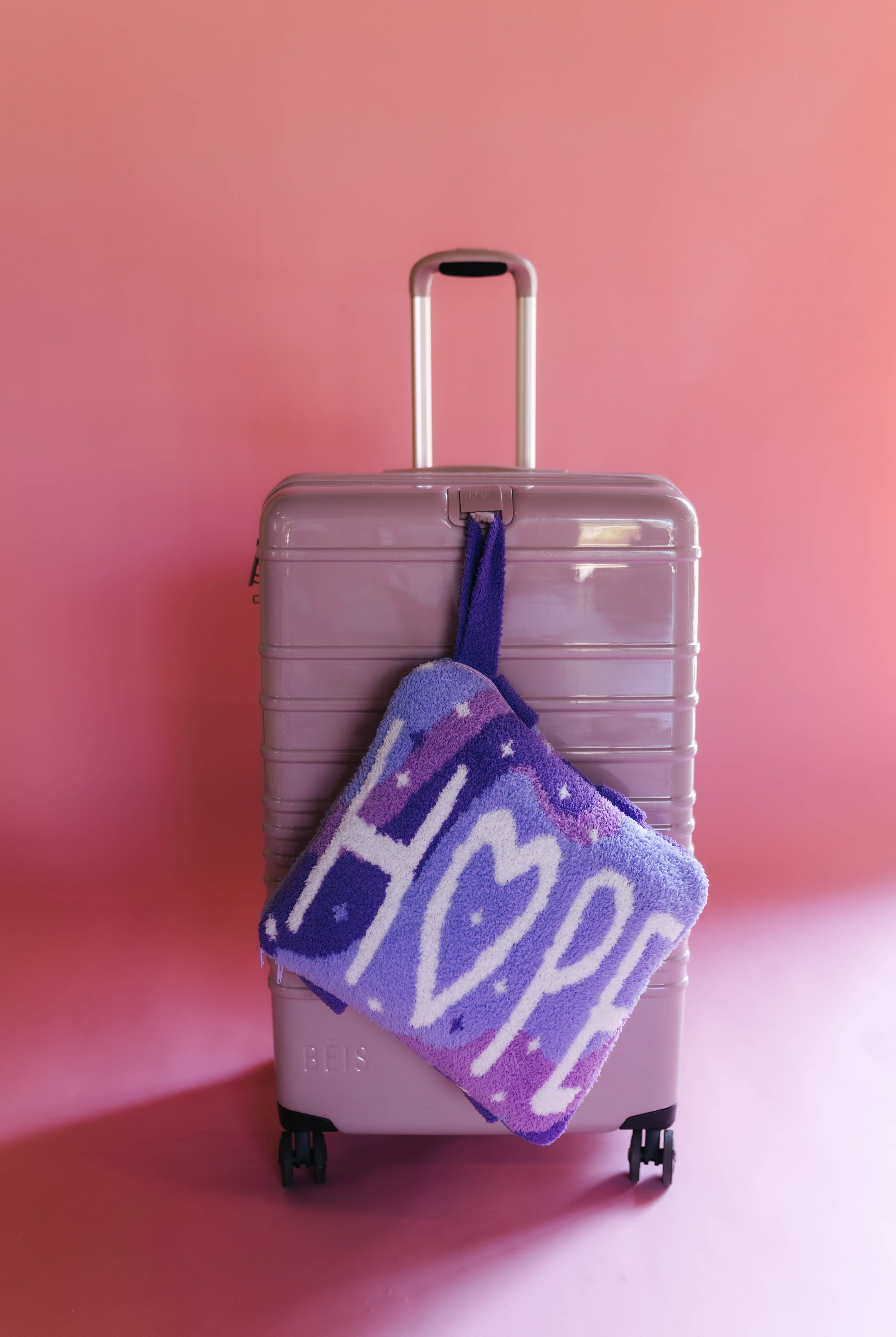 A lavender hard-shell suitcase standing against a pink studio background with the quillow hanging off of the front