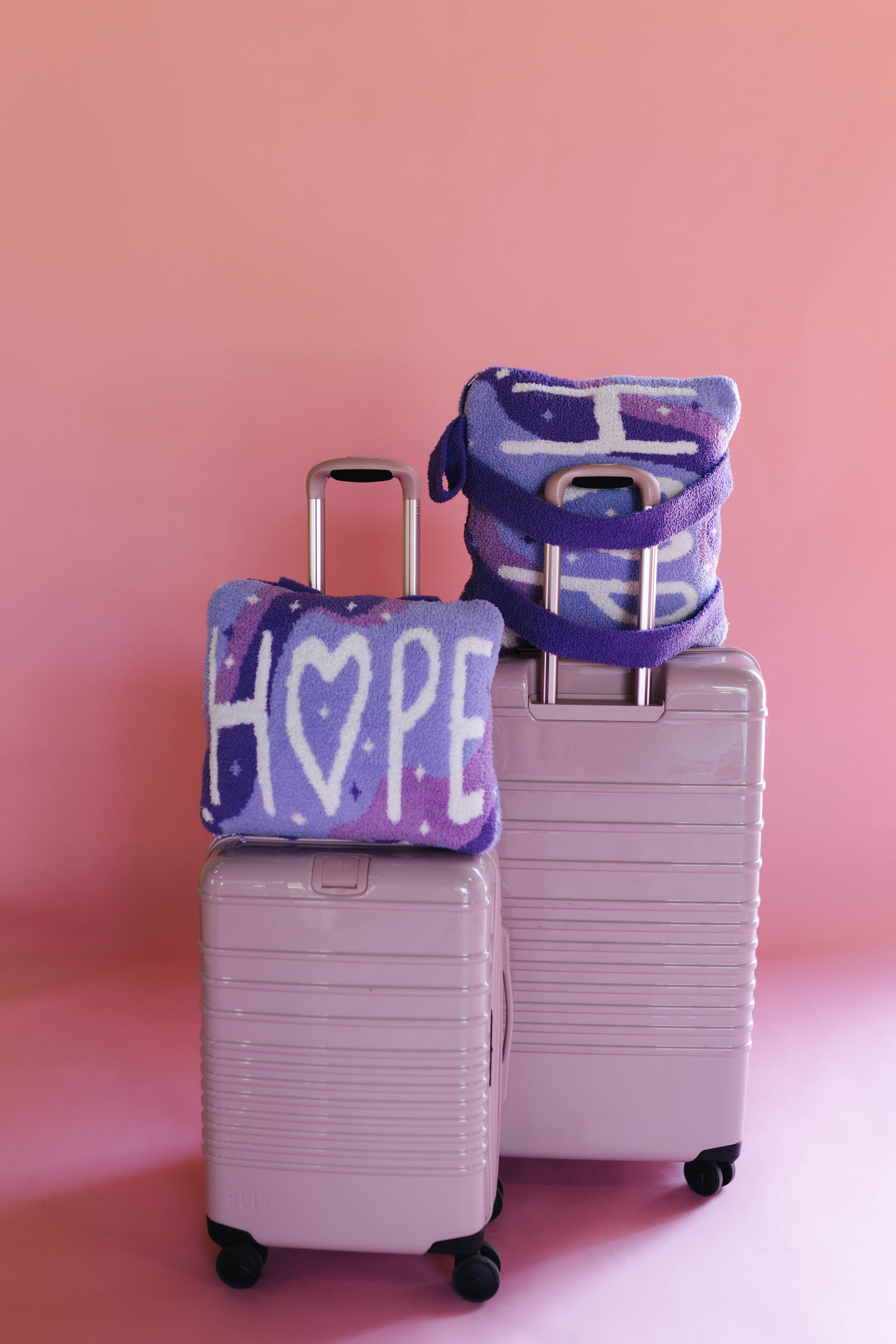 Two pastel pink hard-shell suitcases with matching quillows that say hope