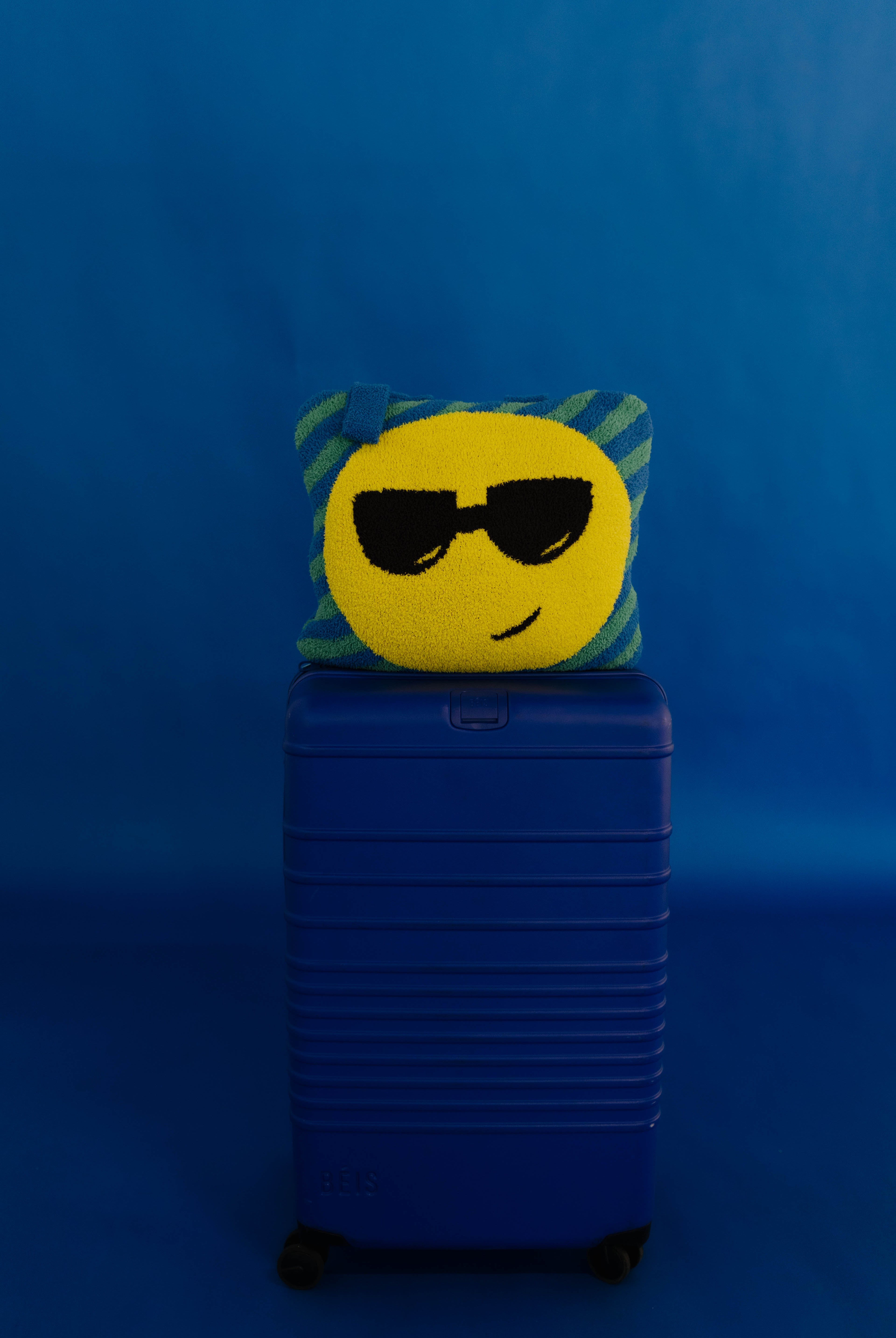 A yellow circular emoji face quillow with black sunglasses and a green striped border, resting on top of a blue hard-shell suitcase against a solid blue background.