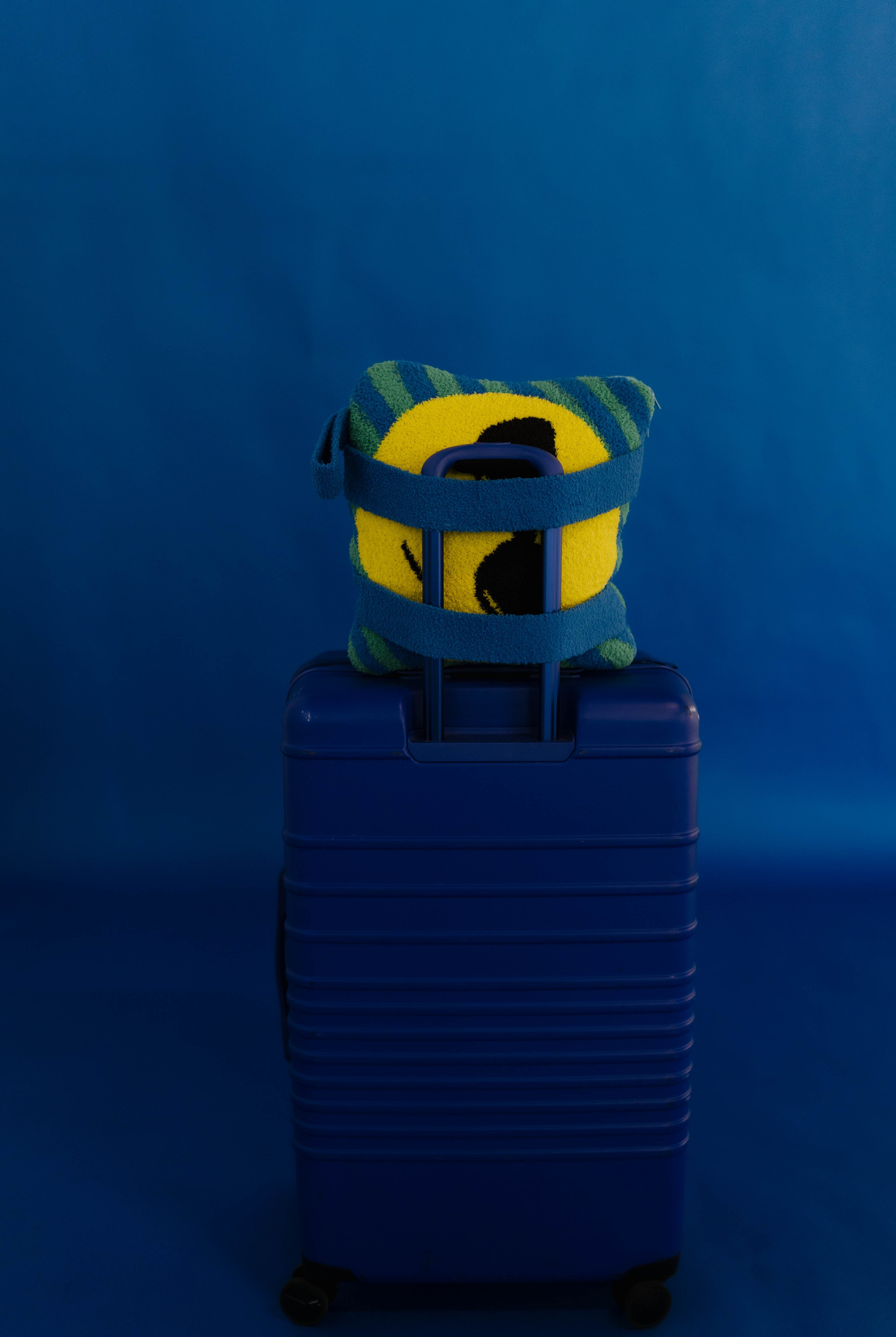 A blue hard-shell rolling suitcase standing upright against a blue studio background, with a yellow and green striped plush quillow tucked into the top handle area.