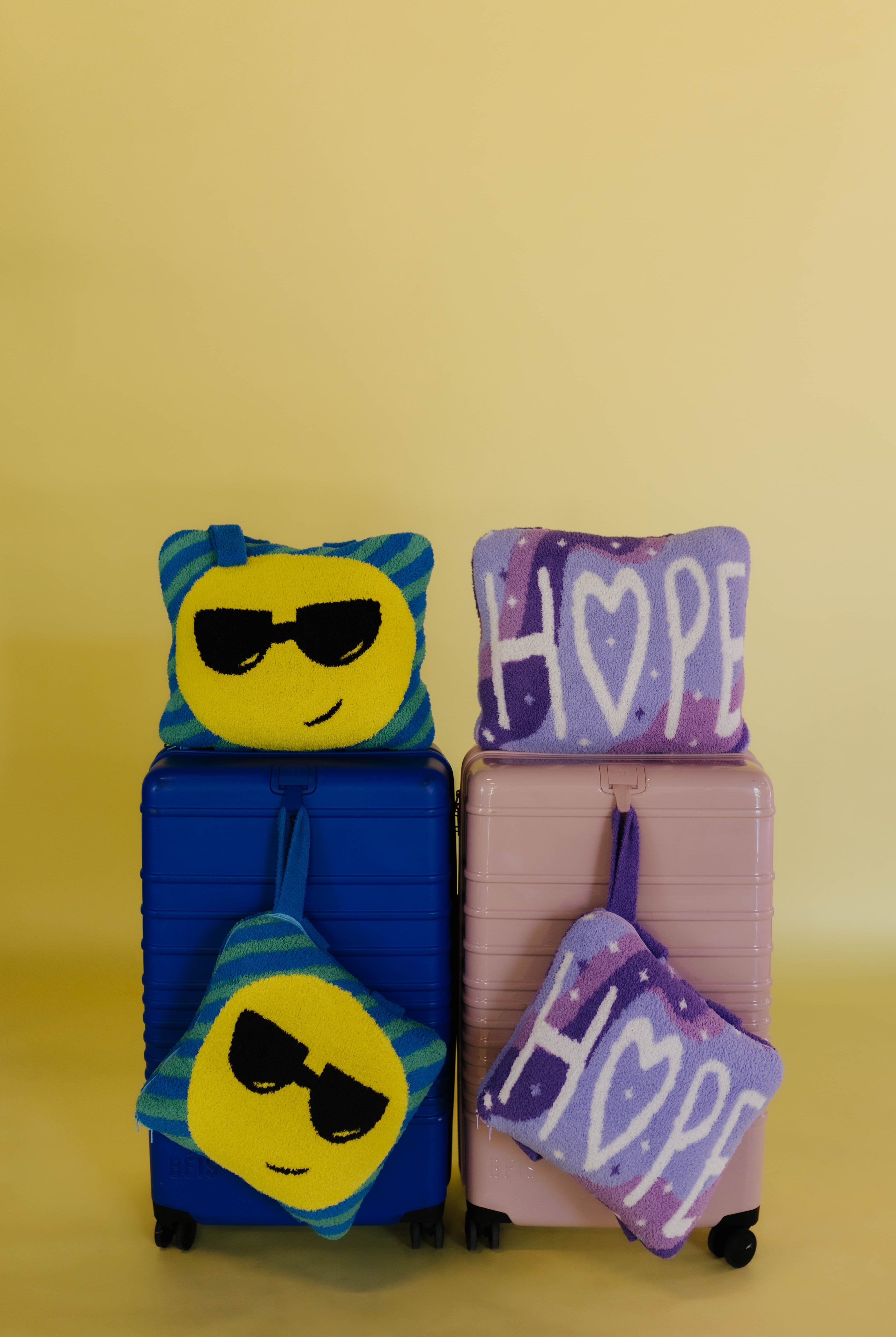 Two hard-shell suitcases, one blue and one pink, each with quillows on them. The blue suitcase has a yellow smiley face with black sunglasses quillow on top and a purple 'HOPE' pouch below.