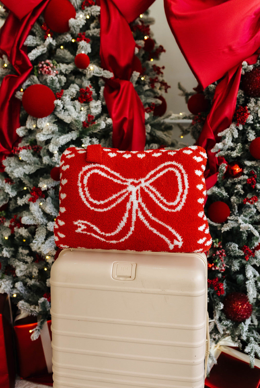 Red quillow with a white bow on a beige suitcase in front of a decorated Christmas tree.