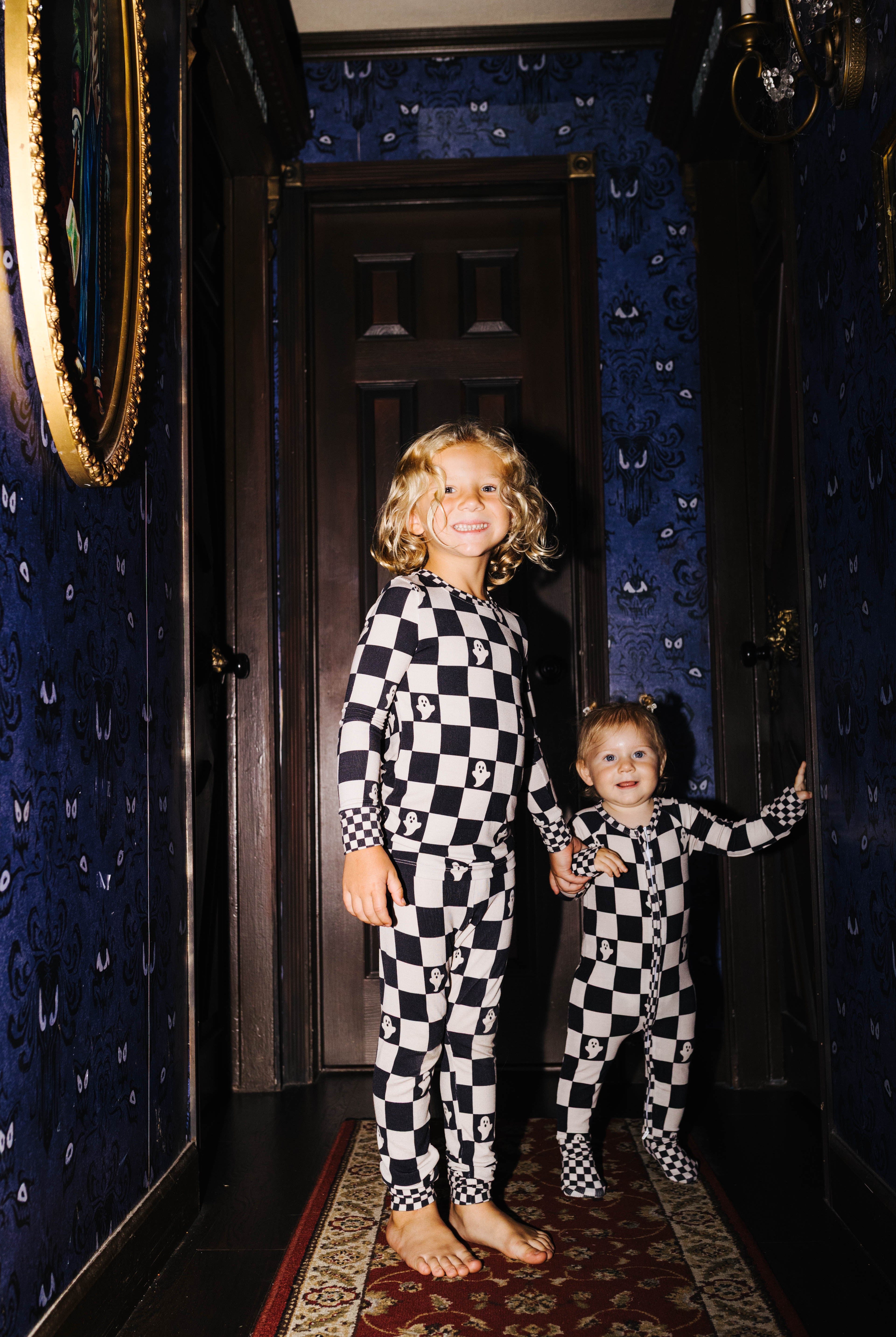 Two children standing in a dark hallway wearing matching black-and-white checkered pajamas. The room has dark blue patterned wallpaper, a dark wooden door behind them, and a red patterned runner rug.