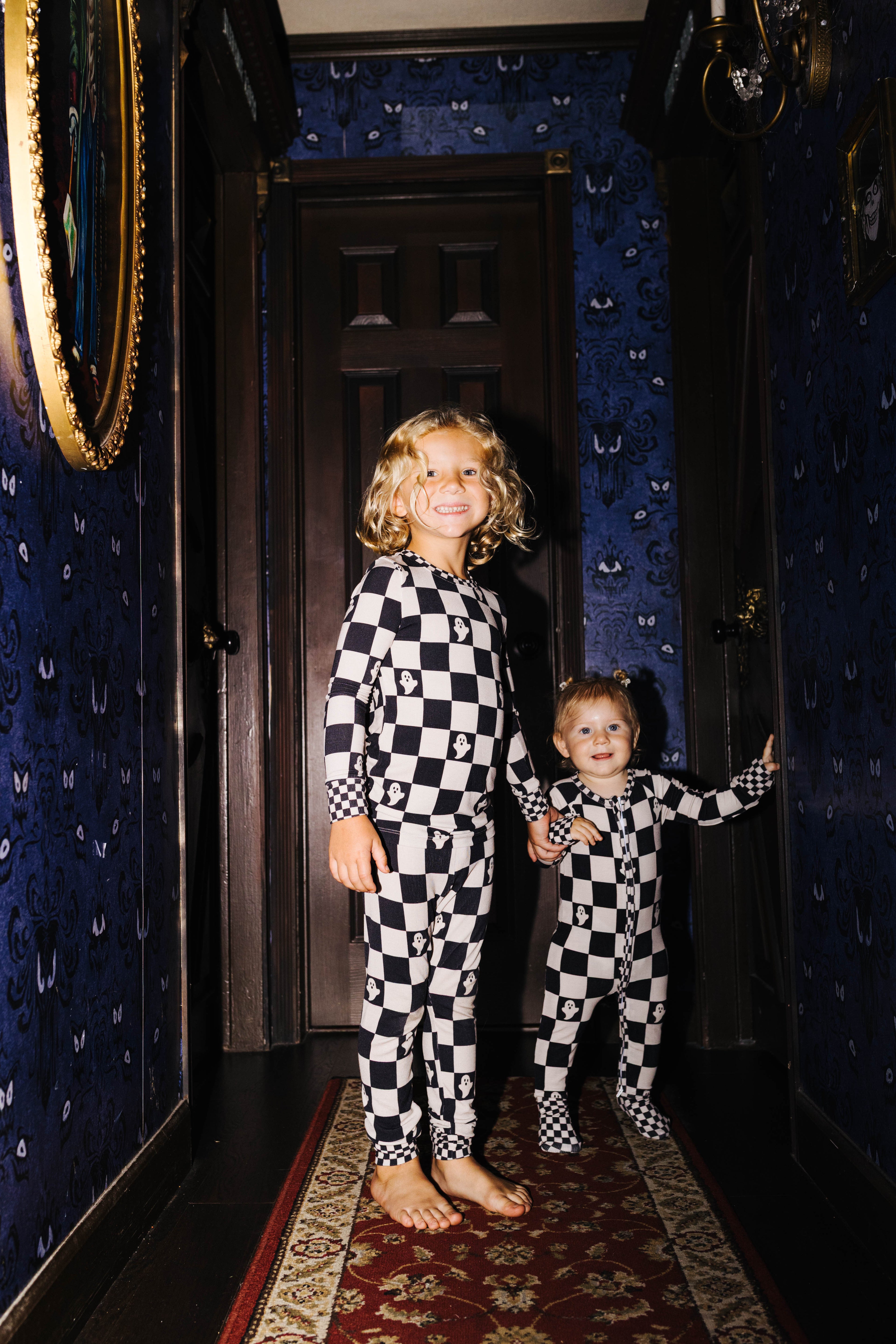 Two children standing in a dark hallway wearing matching black-and-white checkered pajamas. The room has dark blue patterned wallpaper, a dark wooden door behind them, and a red patterned runner rug.