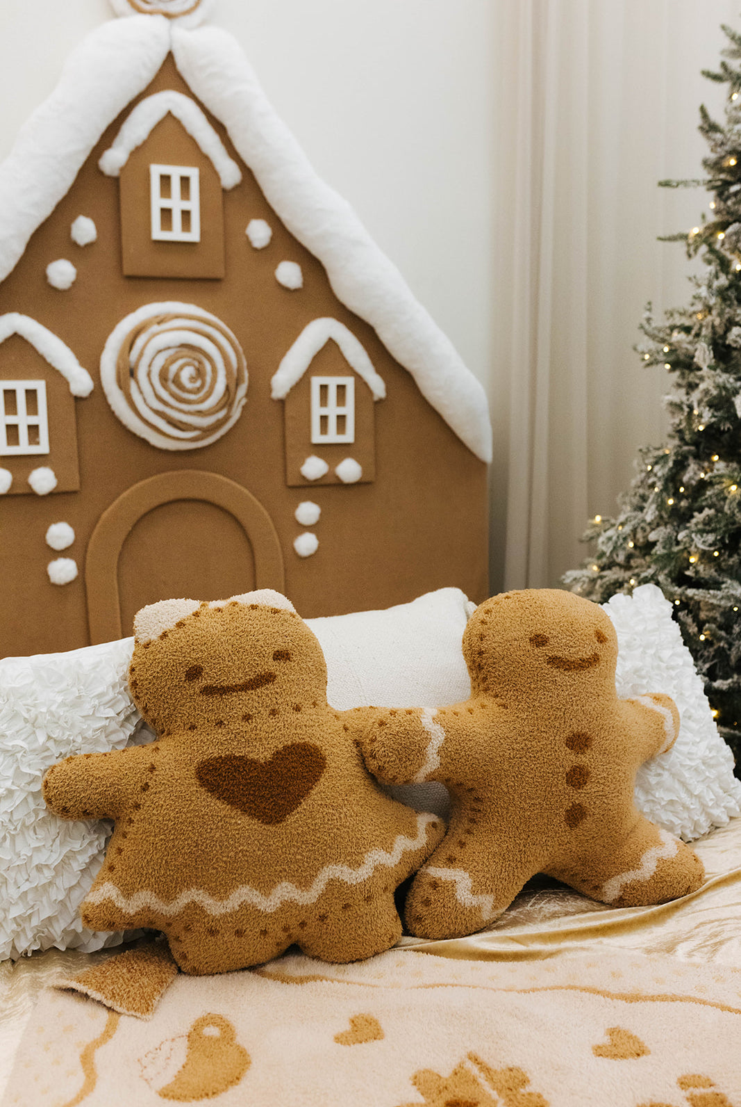 Two plush gingerbread men sitting on a couch with a gingerbread house and Christmas tree in the background.
