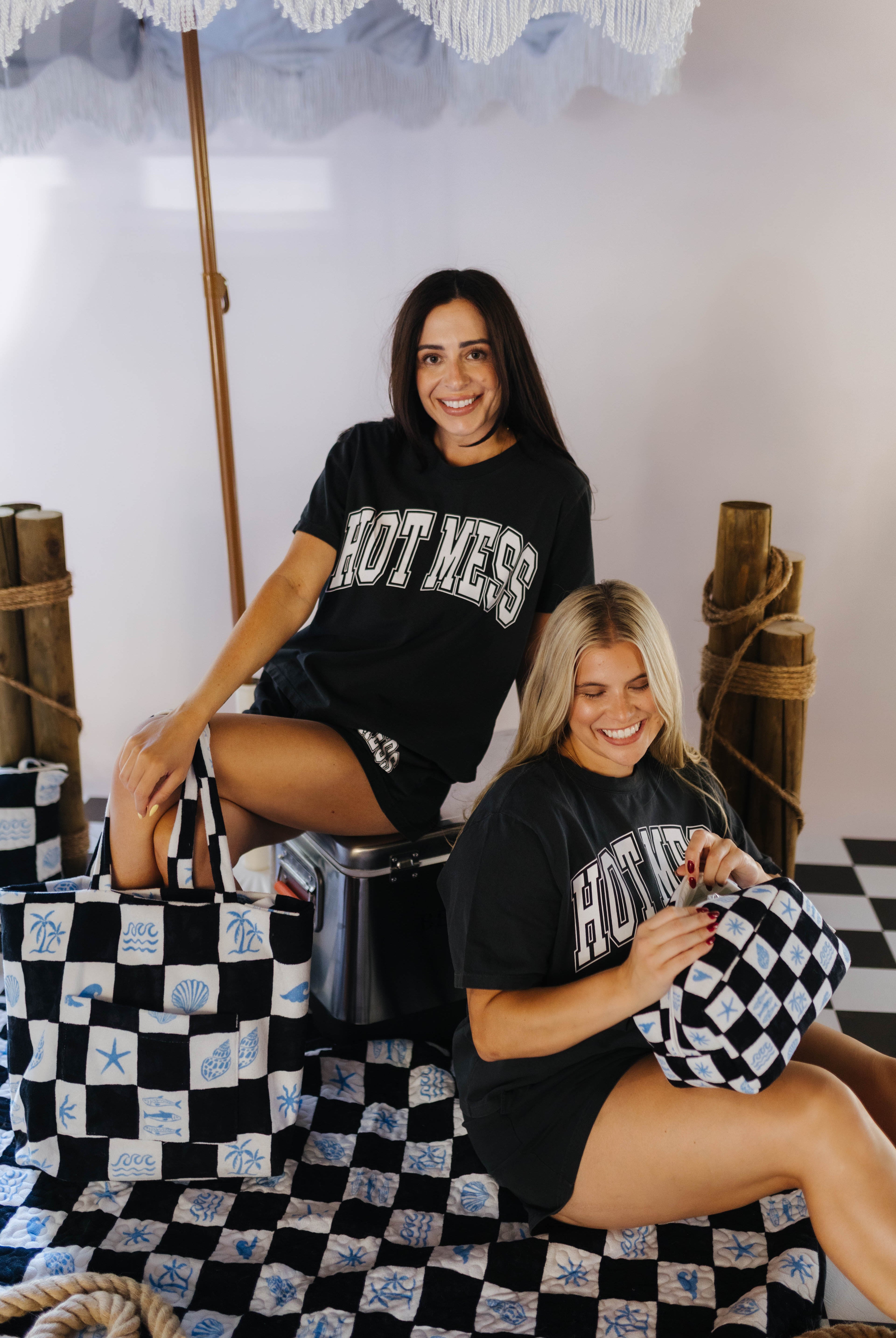 Two women sitting on a checkered floor with black and white bags, wearing matching 'HOTMESS' t-shirts.