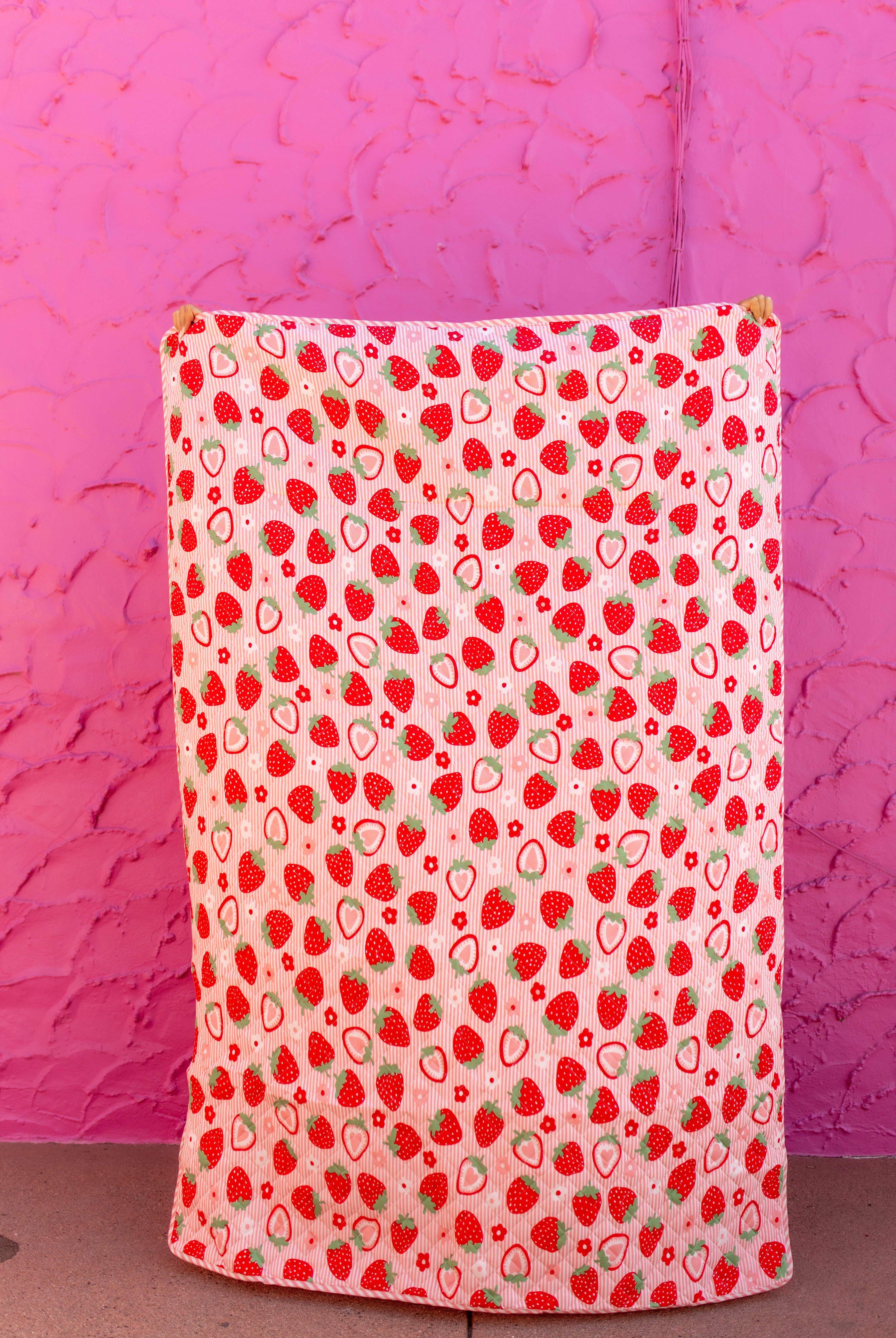 the front of the outdoor quillow with strawberry and subtle pink stripe background held up against a hot pink background