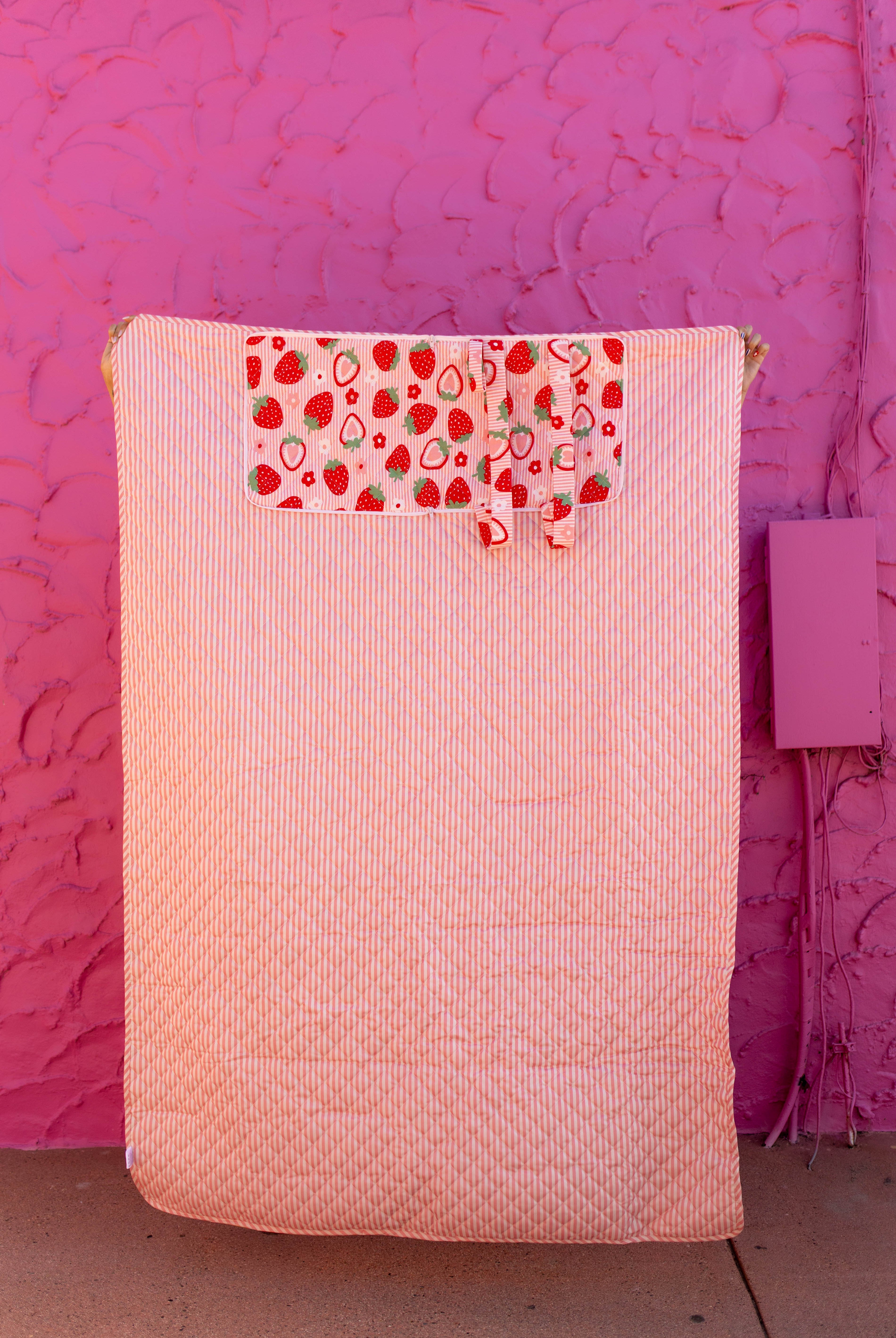 A quilted striped-pink outdoor blanket with a decorative pocket featuring a strawberry and fruit print, set against a textured hot pink wall.