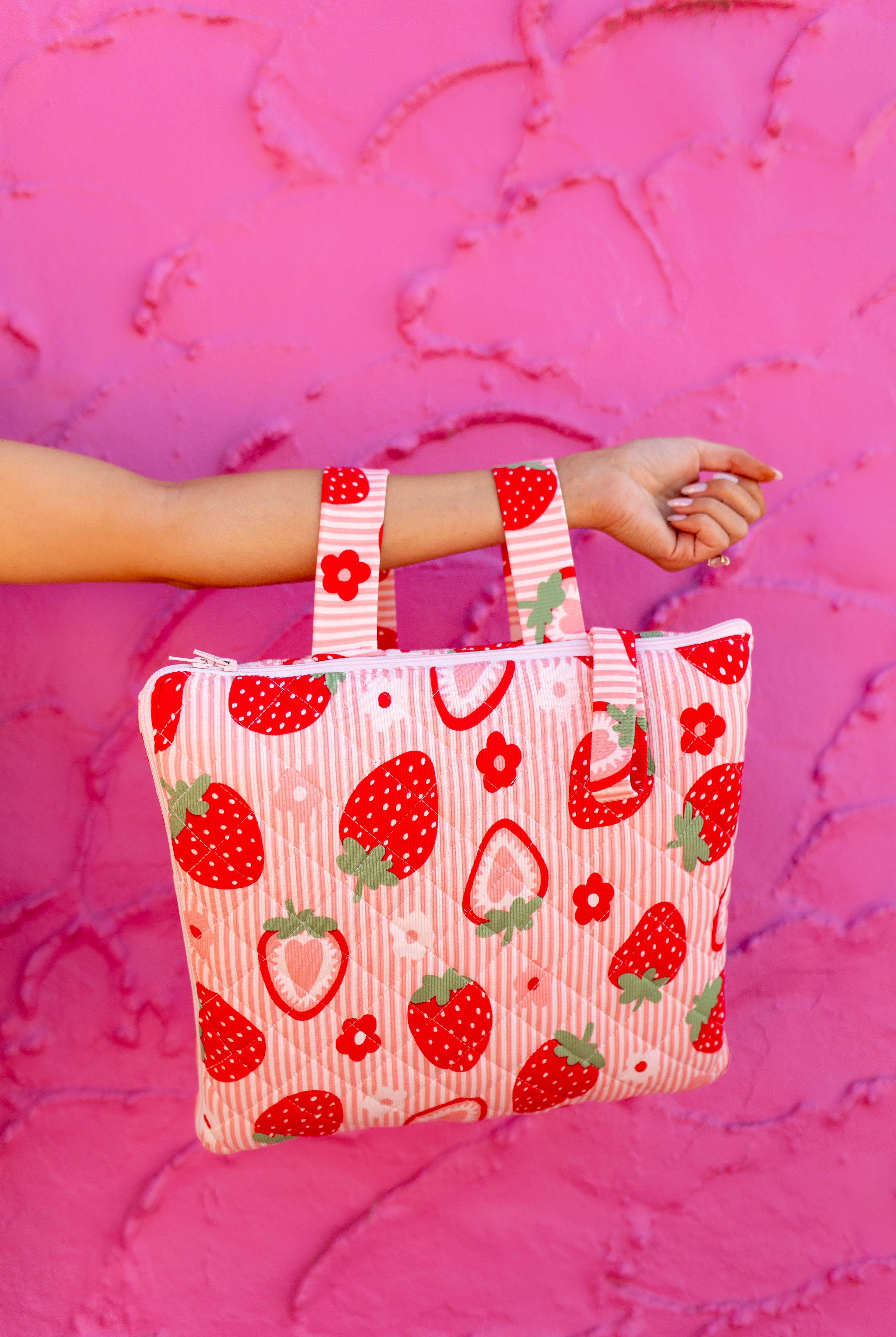 A striped fabric strawberry and flower print quillow held by a person's arm against a textured hot pink background.