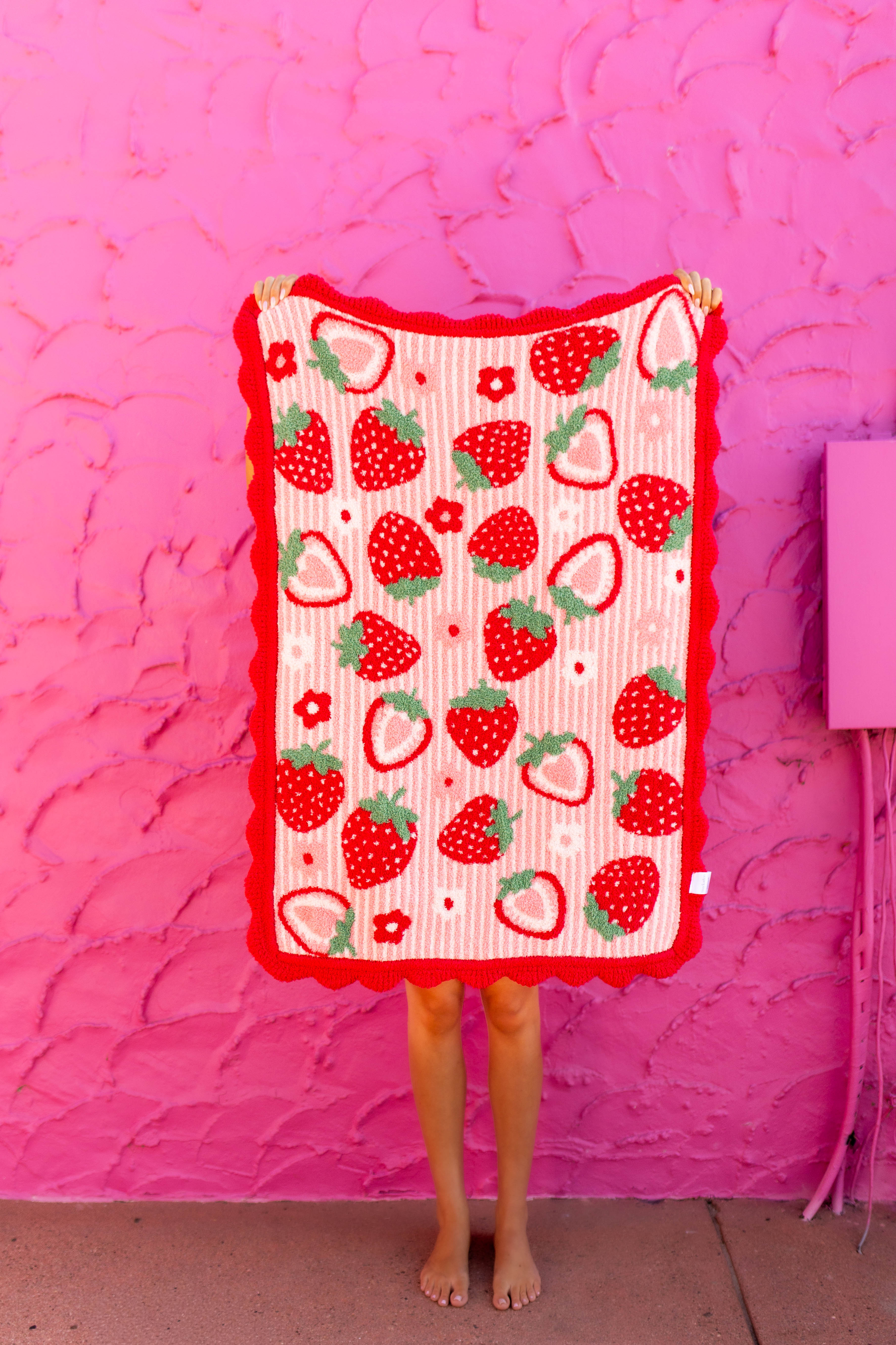 A person holds up a rectangular blanket or throw with a strawberry print pattern, standing barefoot against a bright pink textured wall.
