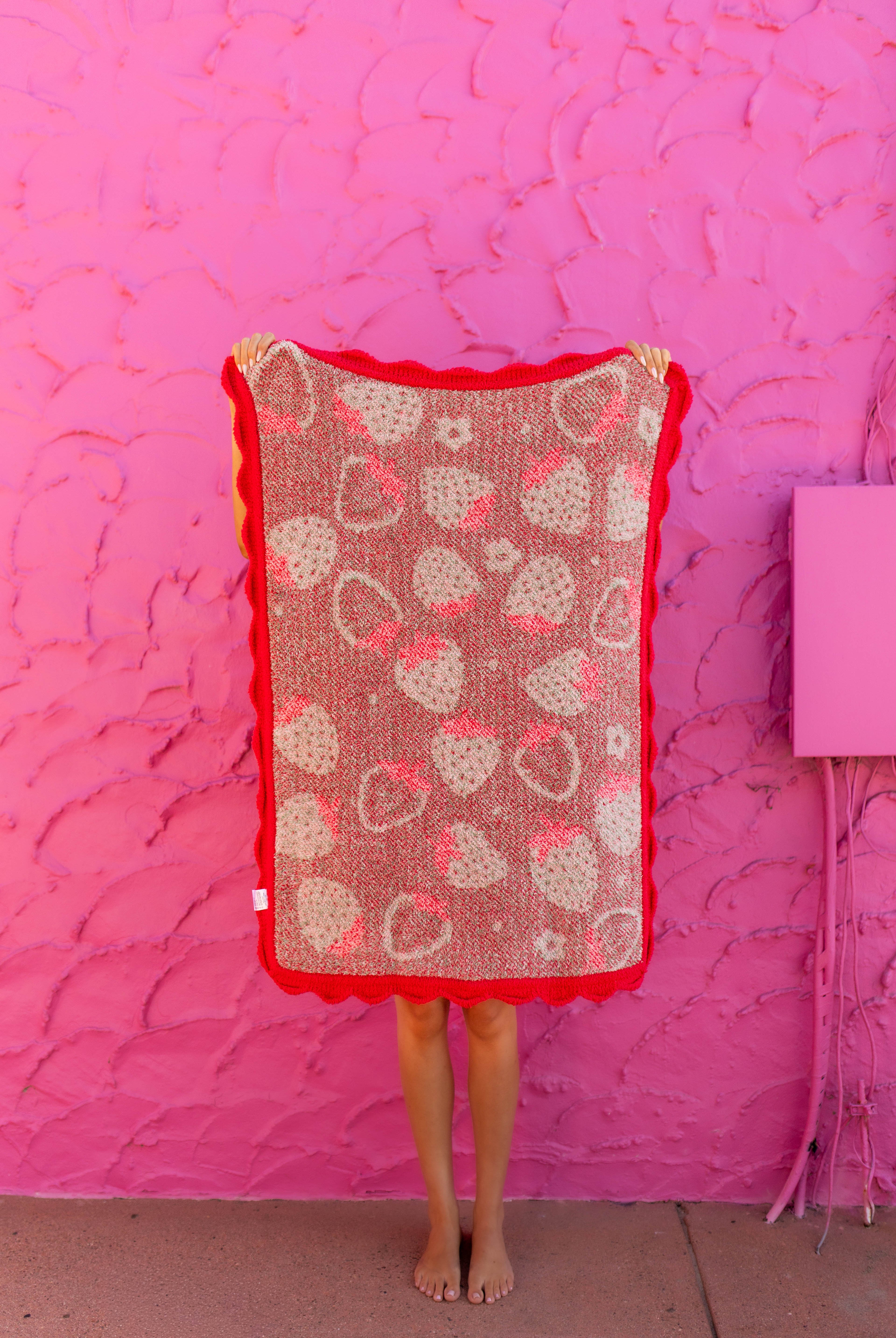 A person standing barefoot in front of a bright pink textured wall, holding up a rectangular crochet blanket with a red border and a pattern of pink and beige hearts.