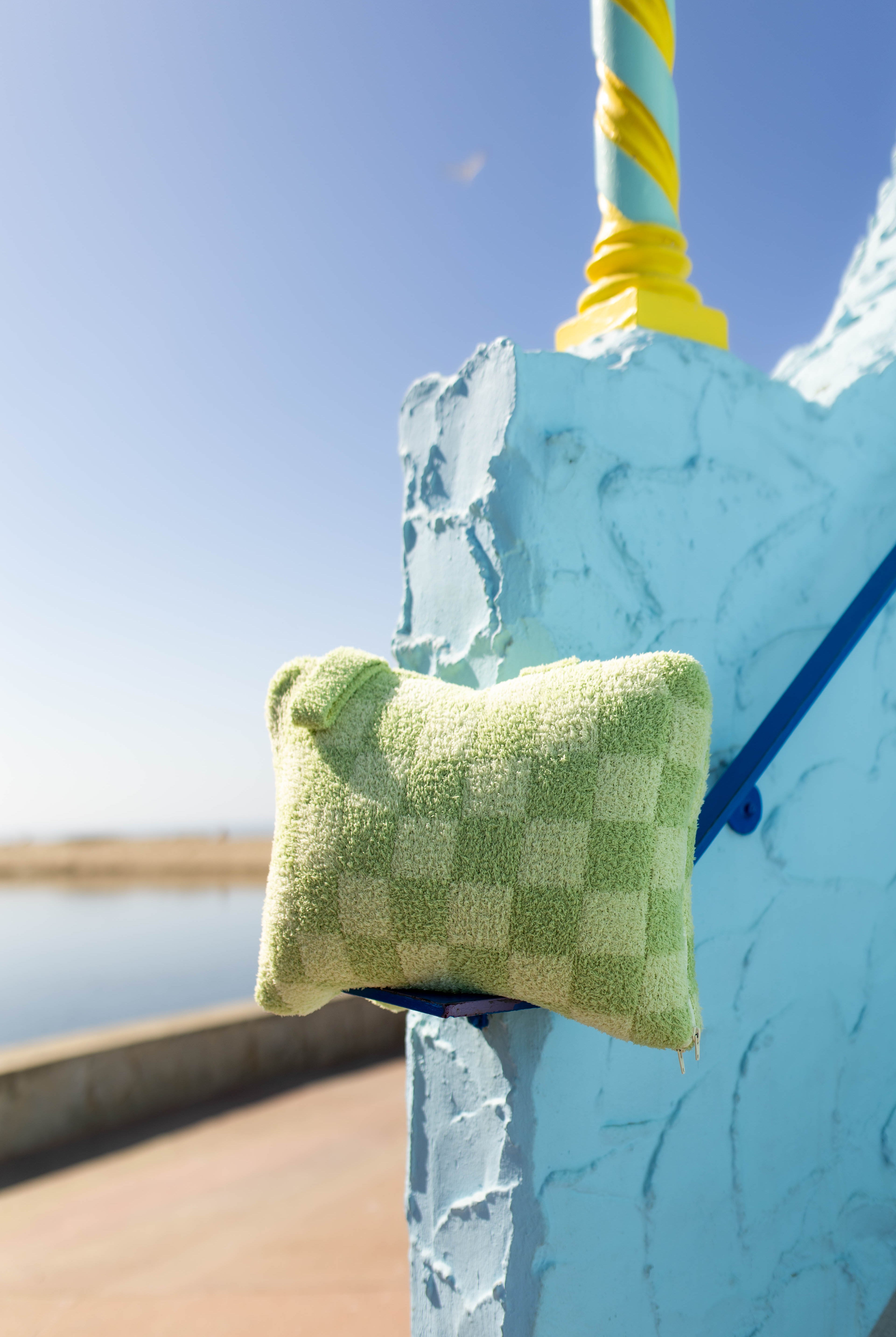 Quillow in a monochromatic check resting on a blue painted structure outdoors near water under a clear blue sky.
