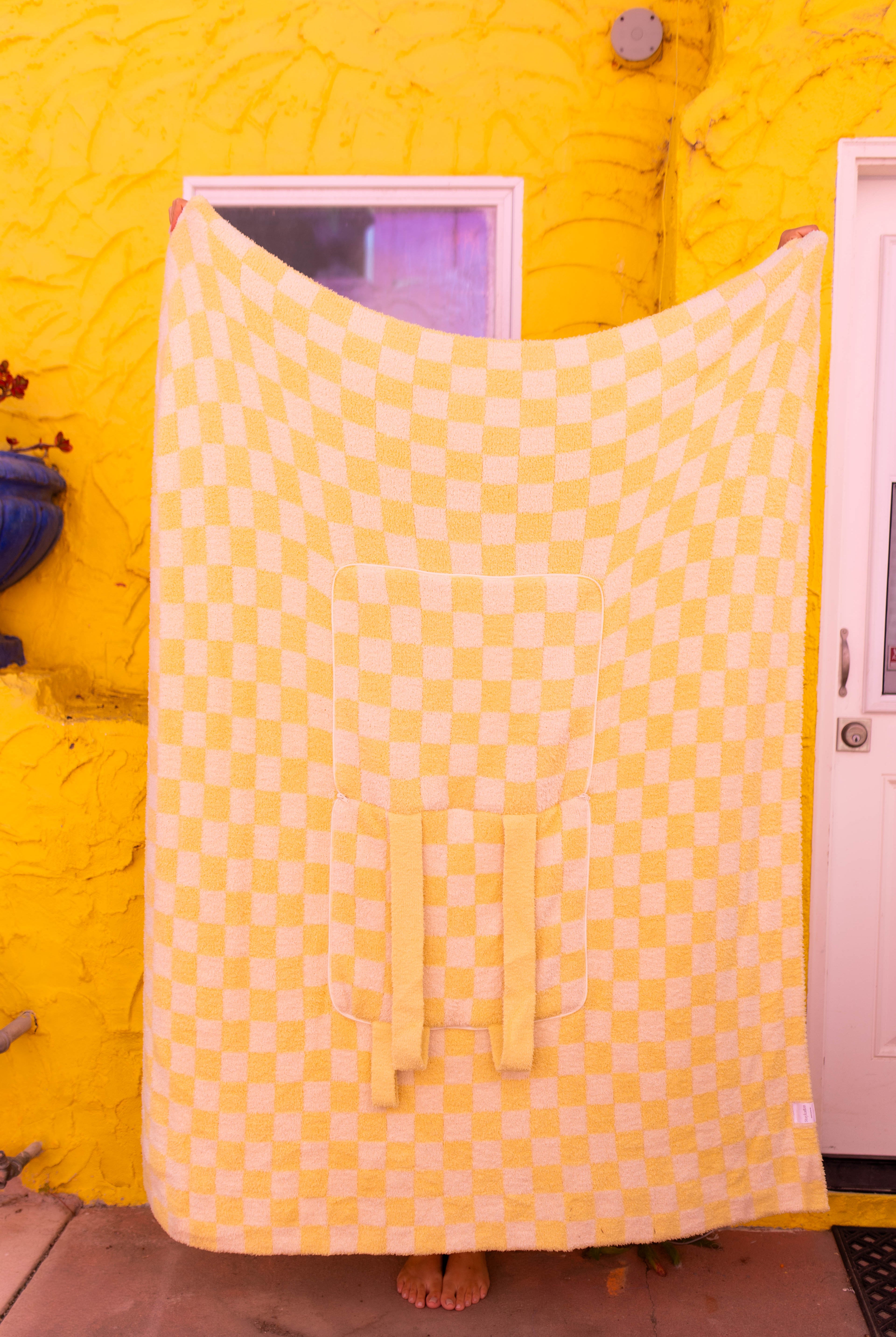 A large checkered yellow quillow is held up by a person standing barefoot in front of a bright yellow stucco wall with a white door.