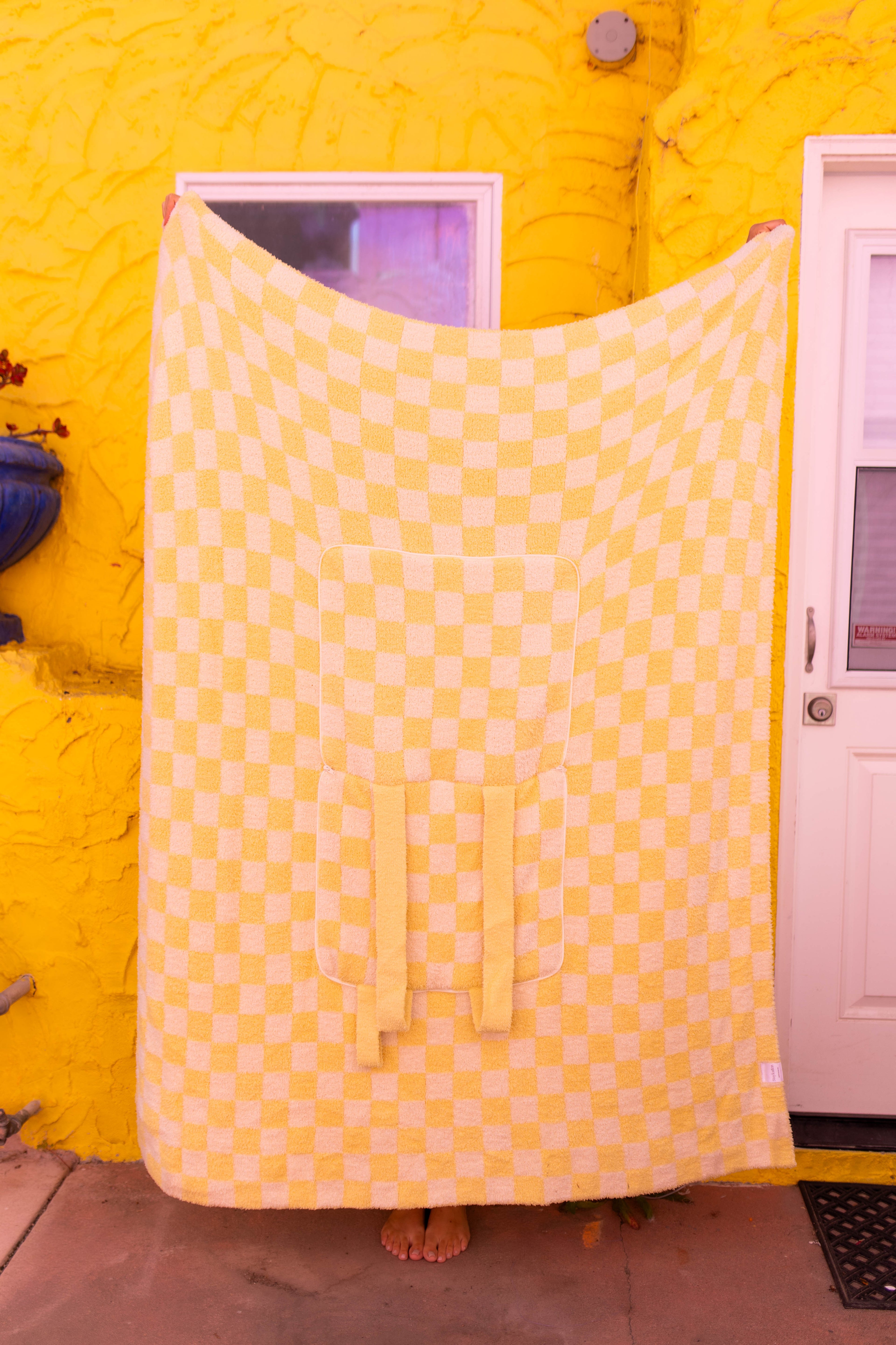 A large checkered yellow quillow is held up by a person standing barefoot in front of a bright yellow stucco wall with a white door.
