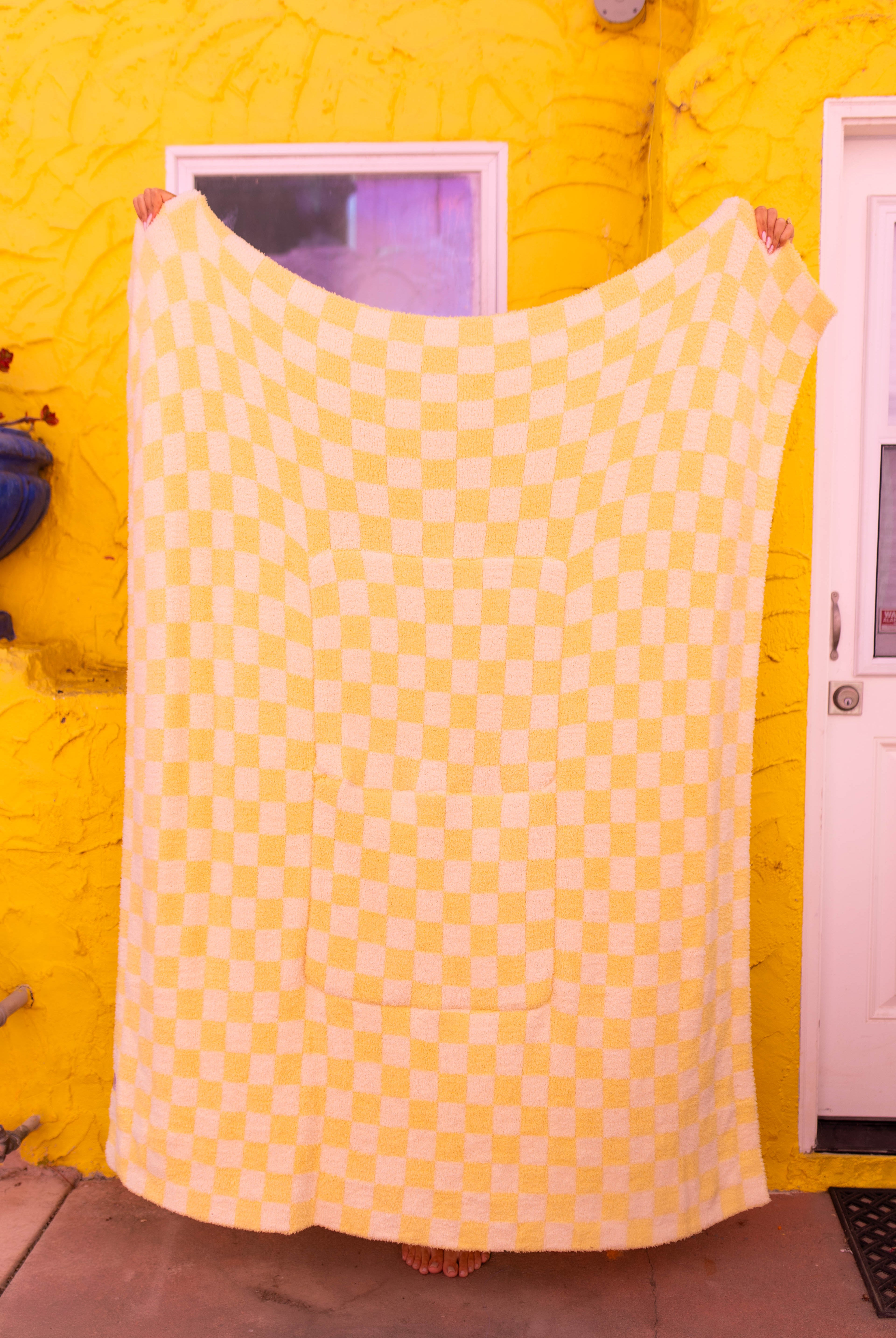 A large yellow checkered quillow is held up by a person in front of a bright yellow wall with a white door visible on the right.