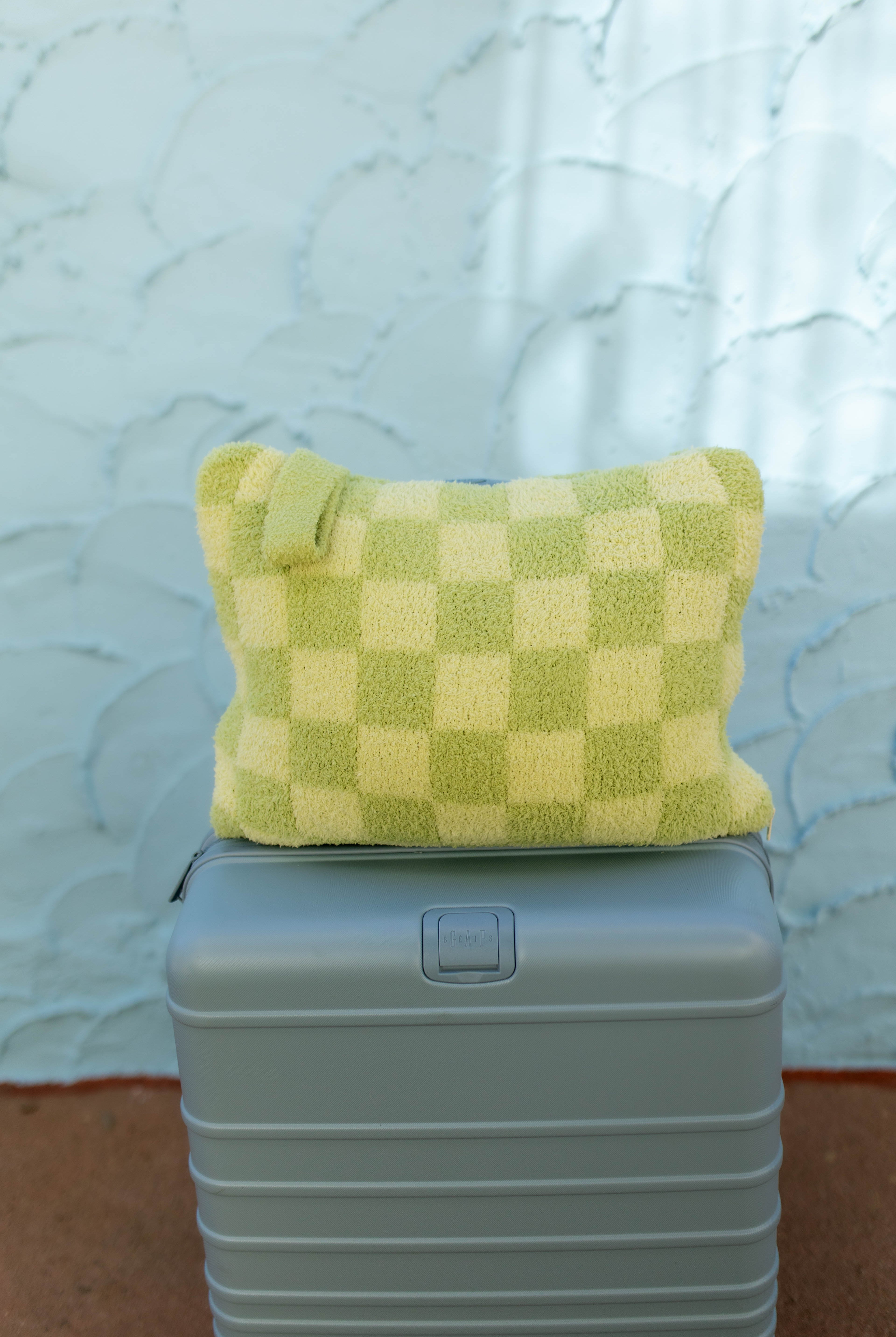 A light green checkered plush quillow with a handle, resting on top of a gray hard-shell suitcase against a textured light blue wall.