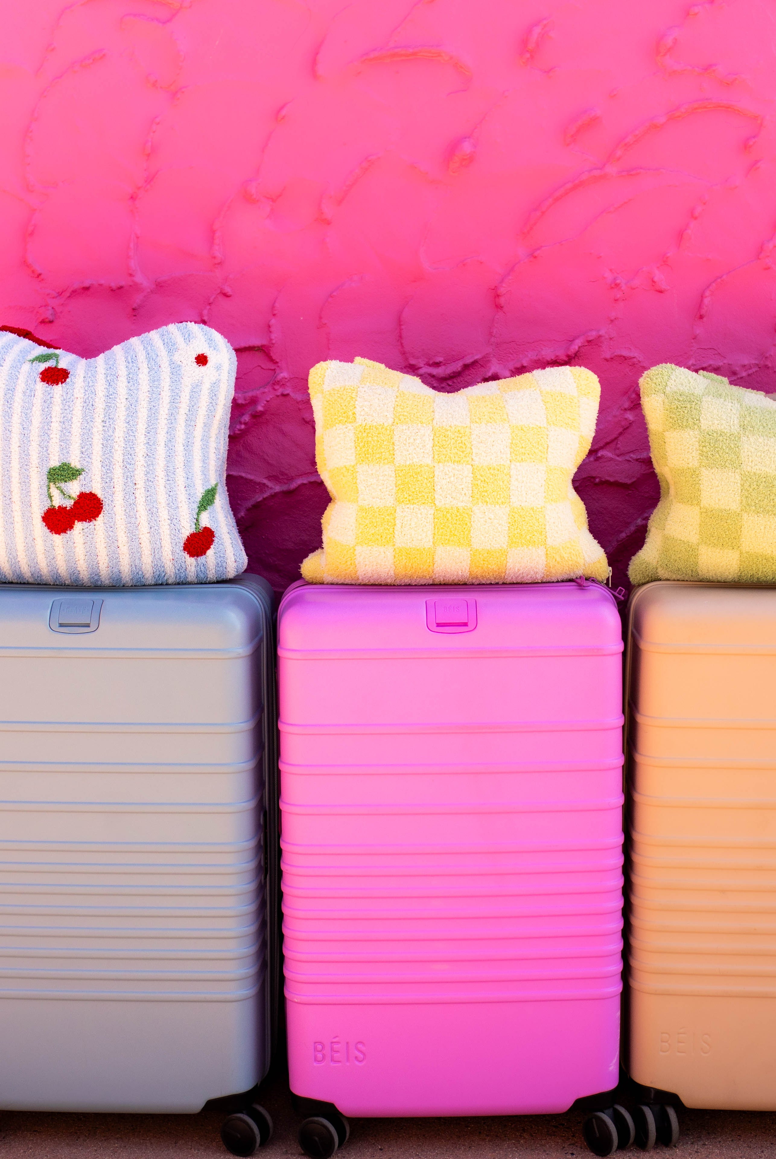 Three hard-shell rolling suitcases in pastel blue, pink, and tan are lined up against a bright pink textured wall, each topped with a quillow.