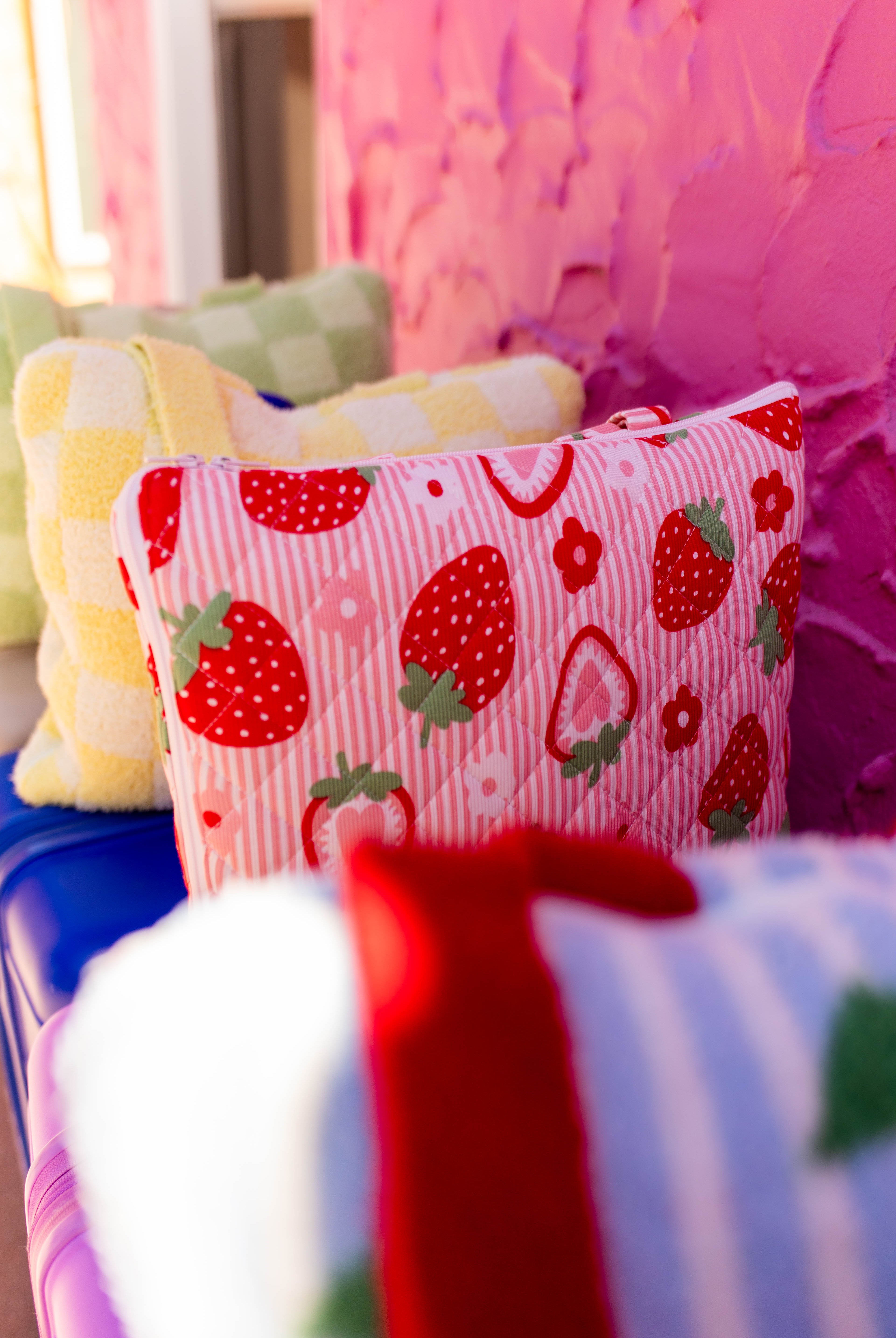 A close-up of a pink striped pillow with a red strawberry pattern, resting on a blue surface in front of a textured pink wall.