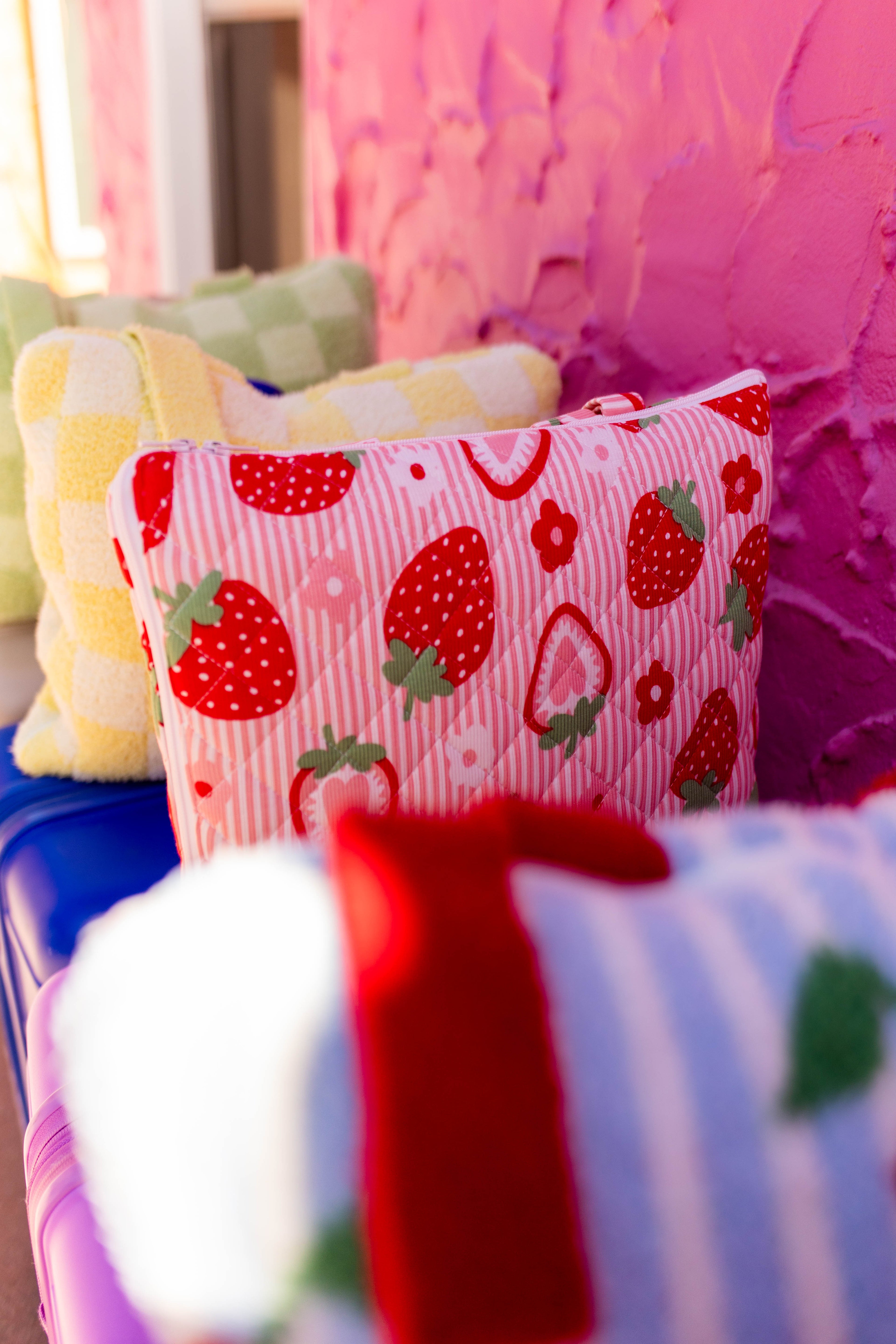 A close-up of a pink striped pillow with a red strawberry pattern, resting on a blue surface in front of a textured pink wall.