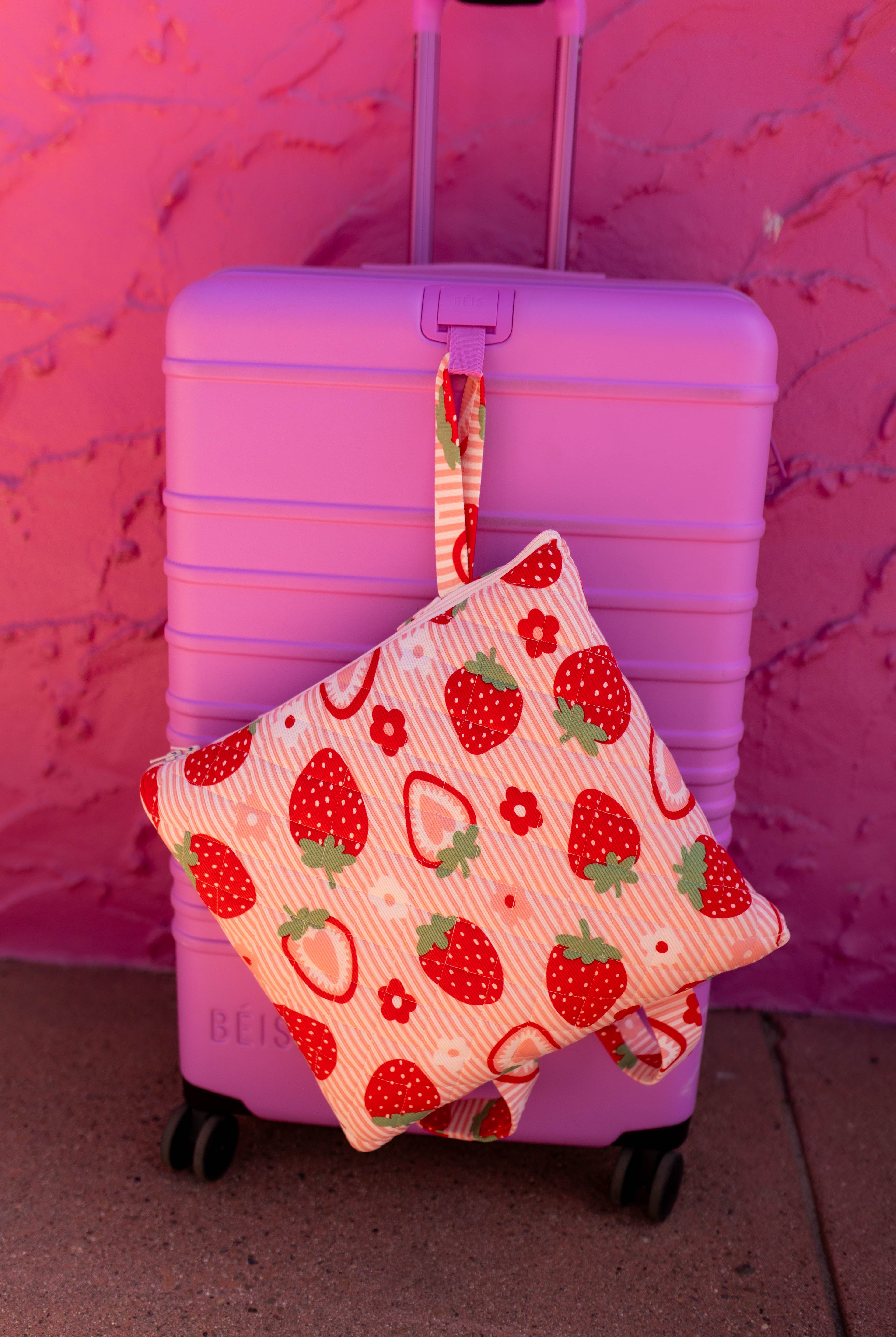 Our outdoor quillow with a strawberry and hanging from a pink suitcase against a pink textured wall.