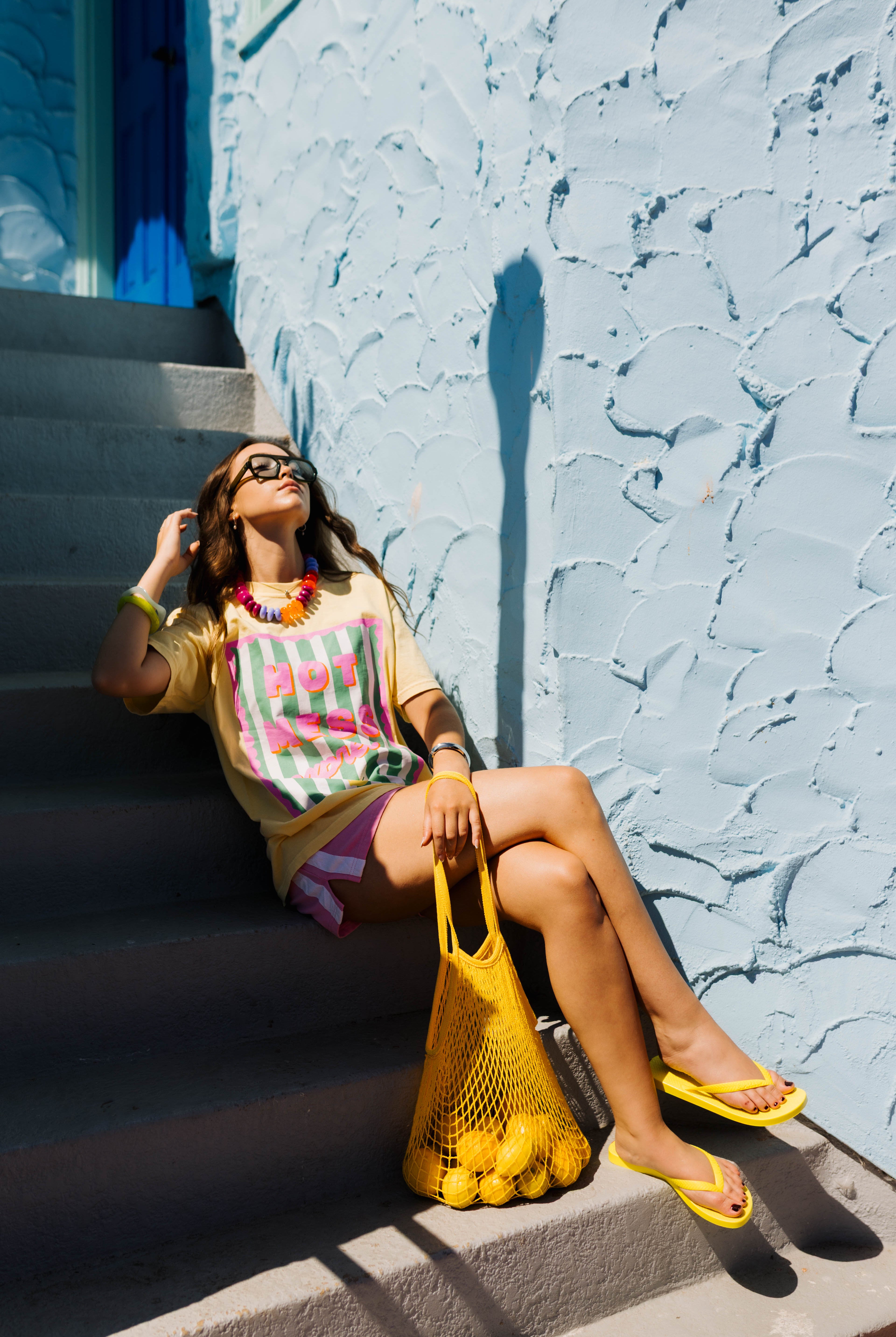 A woman sits on outdoor concrete stairs against a textured light blue wall, wearing a colorful striped graphic T-shirt, pink shorts, yellow flip-flops, and holding a yellow mesh bag of lemons.