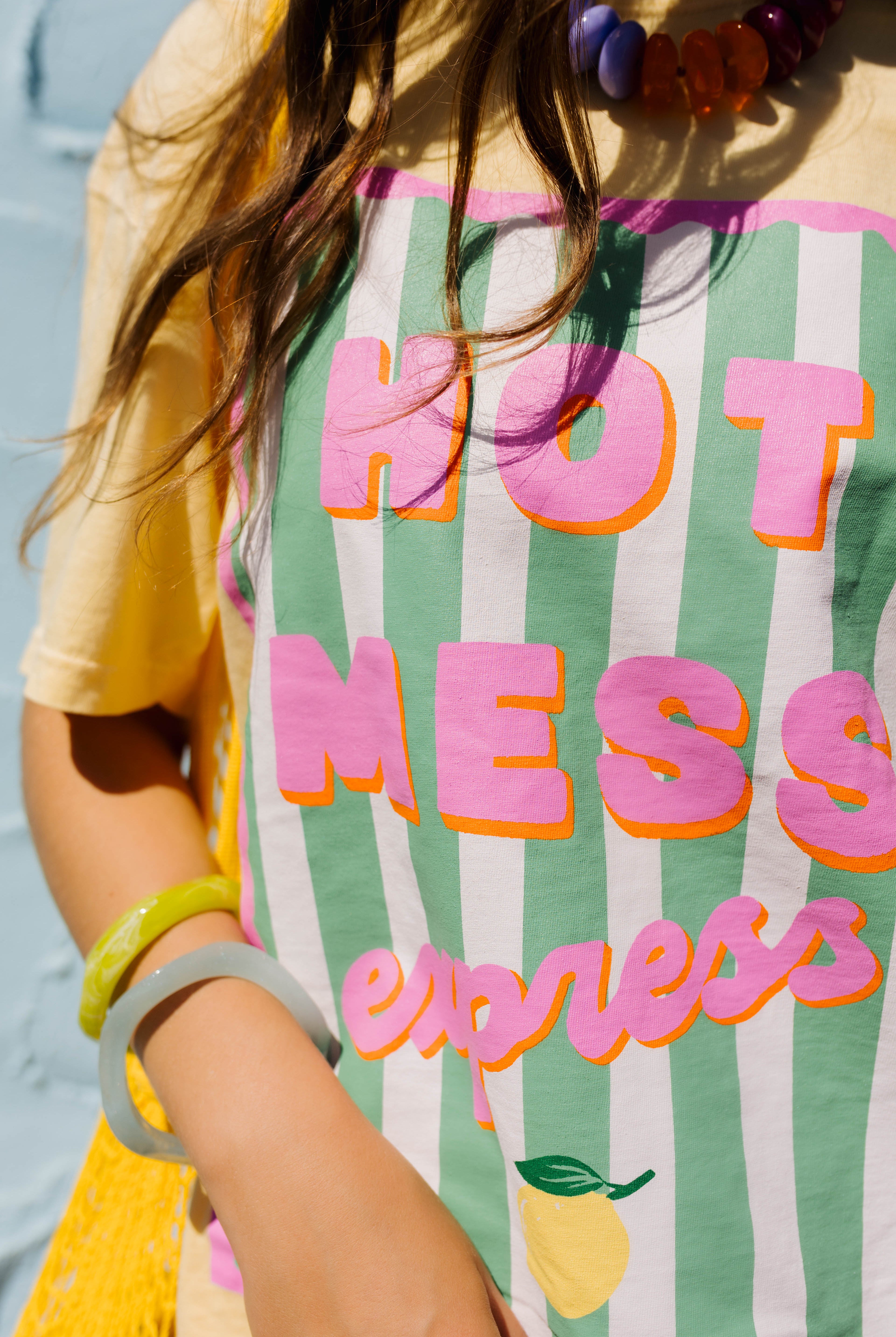 A close-up of a person wearing a yellow t-shirt with a green and white striped graphic that reads 'HOT MESS express' in pink and orange lettering, along with a yellow knit bag and colorful bracelets.