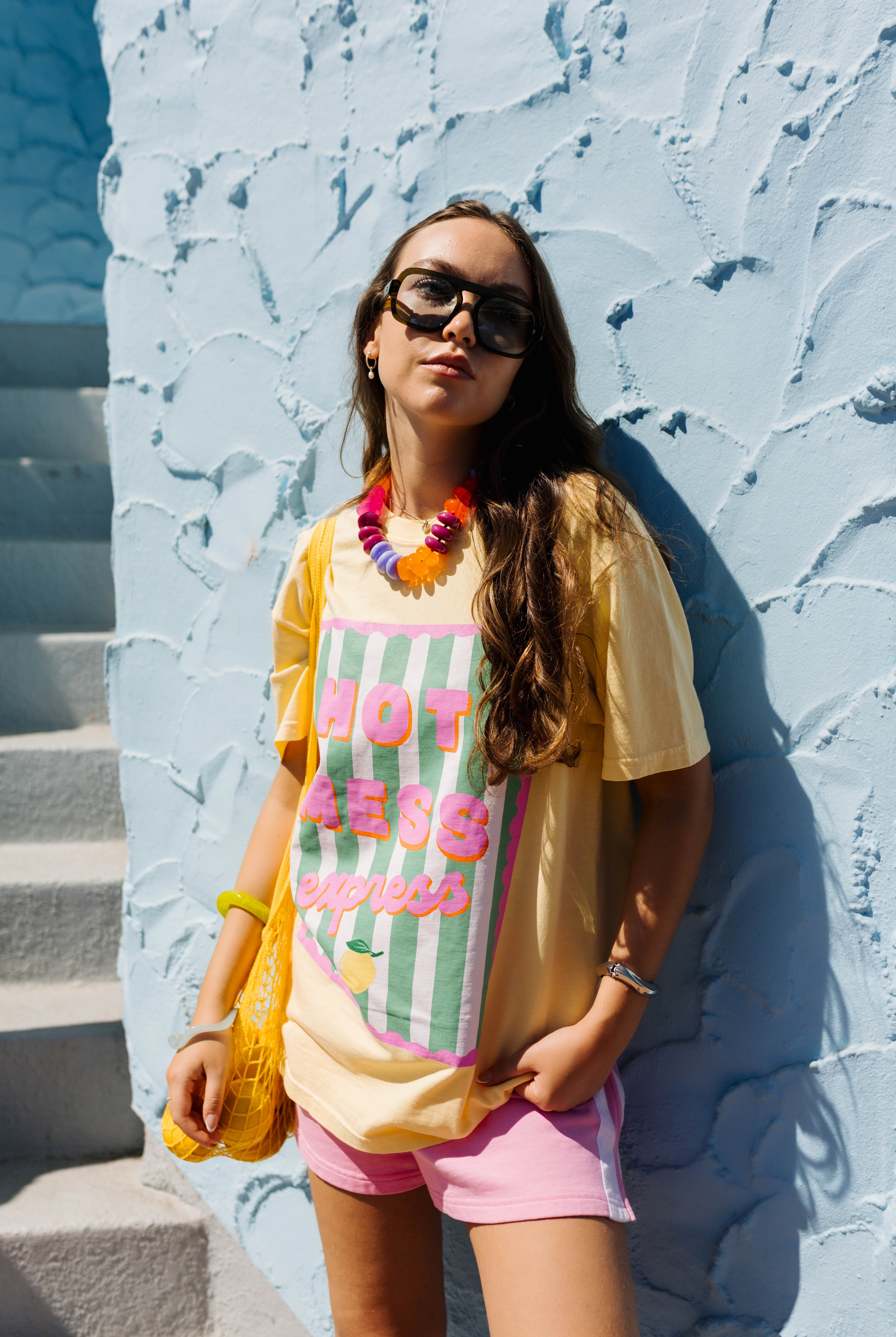 A woman wearing a yellow graphic t-shirt with pink and green text, pink shorts, and a colorful beaded necklace, posing against a textured light blue wall.
