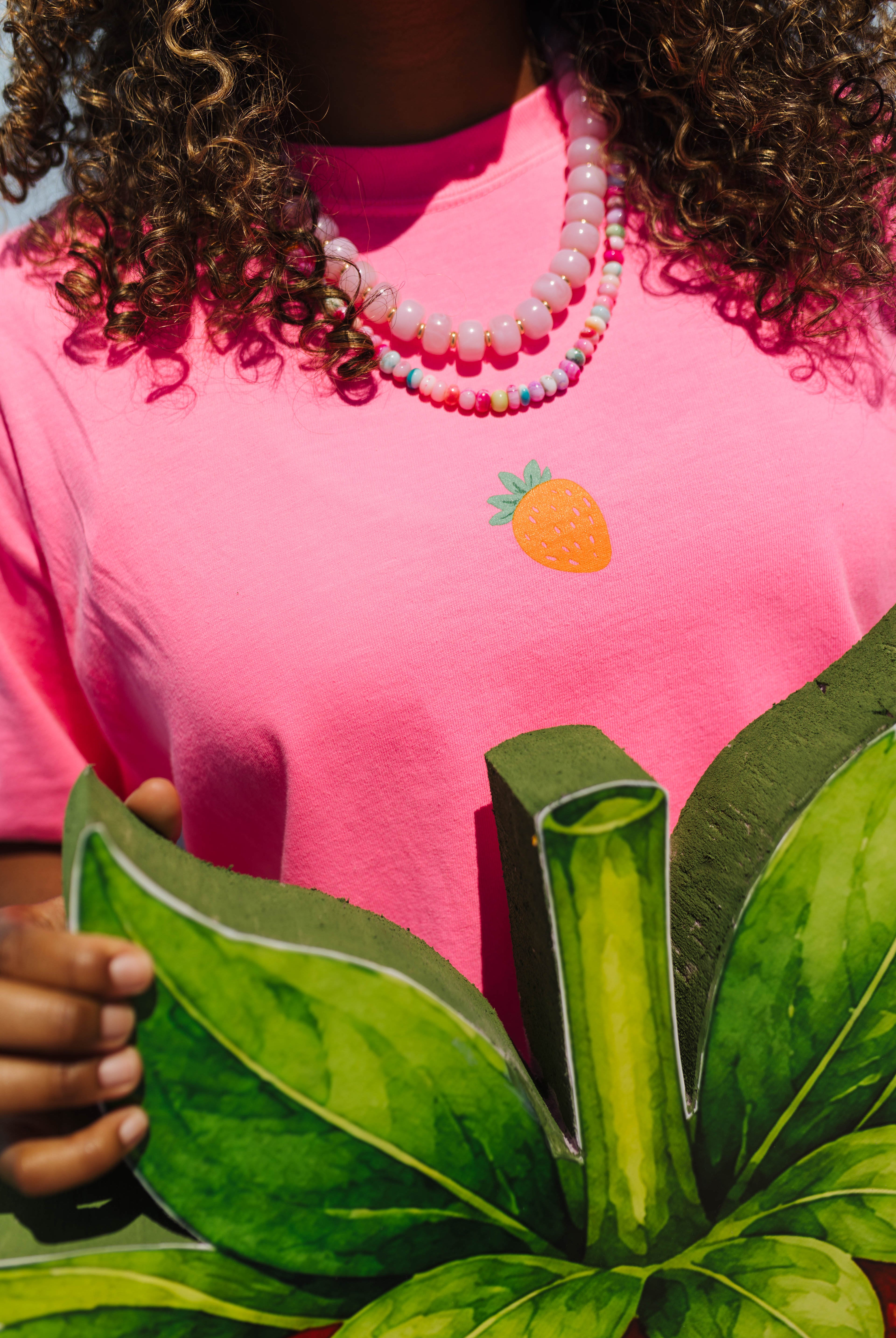 A close-up of a person wearing a bright pink t-shirt with a small embroidered strawberry graphic on the chest. The person is also wearing a layered beaded necklace and holding a large green leafy cutout prop.