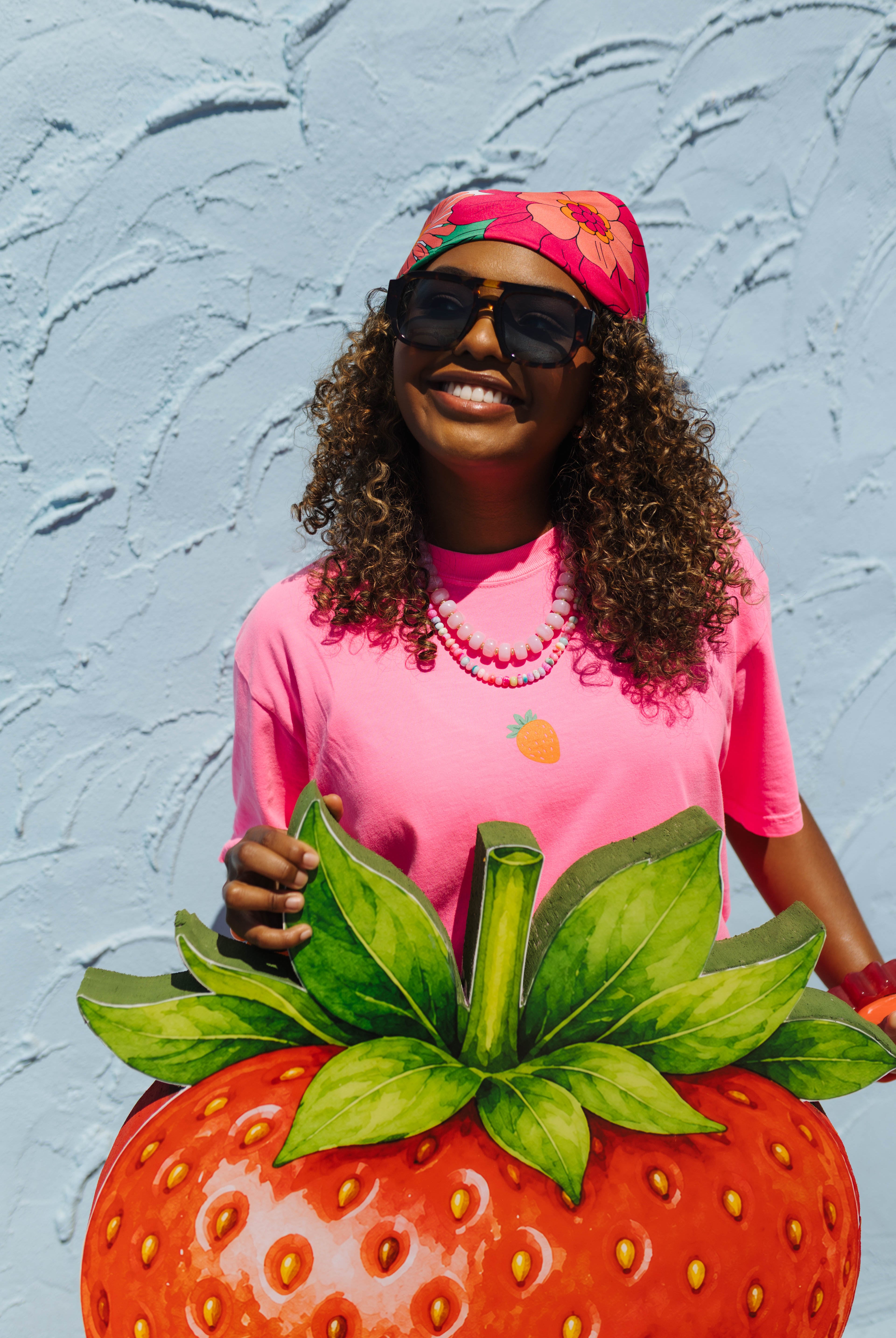A woman wearing a pink t-shirt with a pineapple graphic, a pink floral headband, and sunglasses, holding a giant strawberry sculpture against a light blue textured wall.