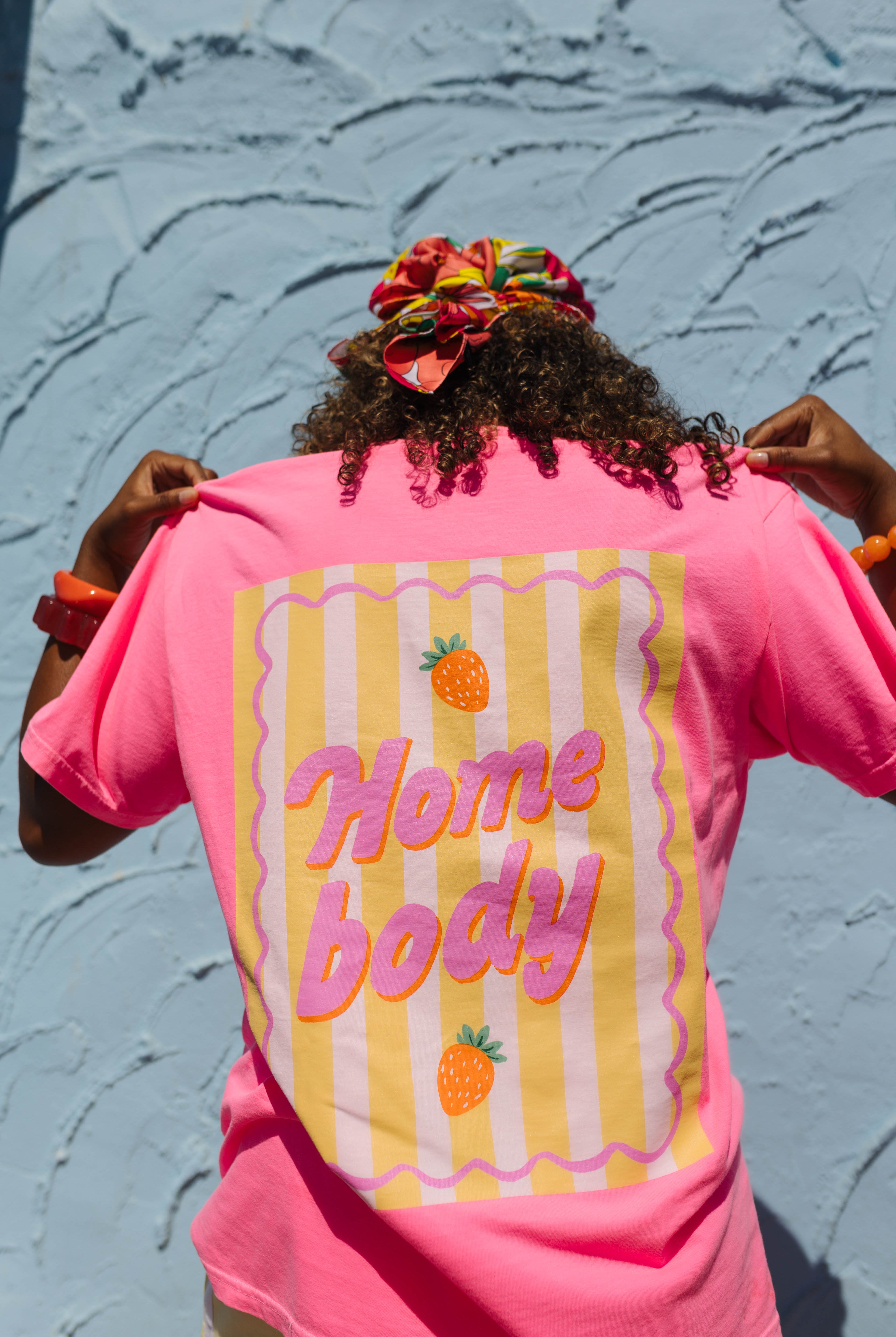 A person viewed from behind wearing a bright pink t-shirt with a retro graphic on the back. The graphic features yellow and white vertical stripes, wavy pink borders, and the words 'Home body' in pink bubble letters, accented with small orange pineapple illustrations.