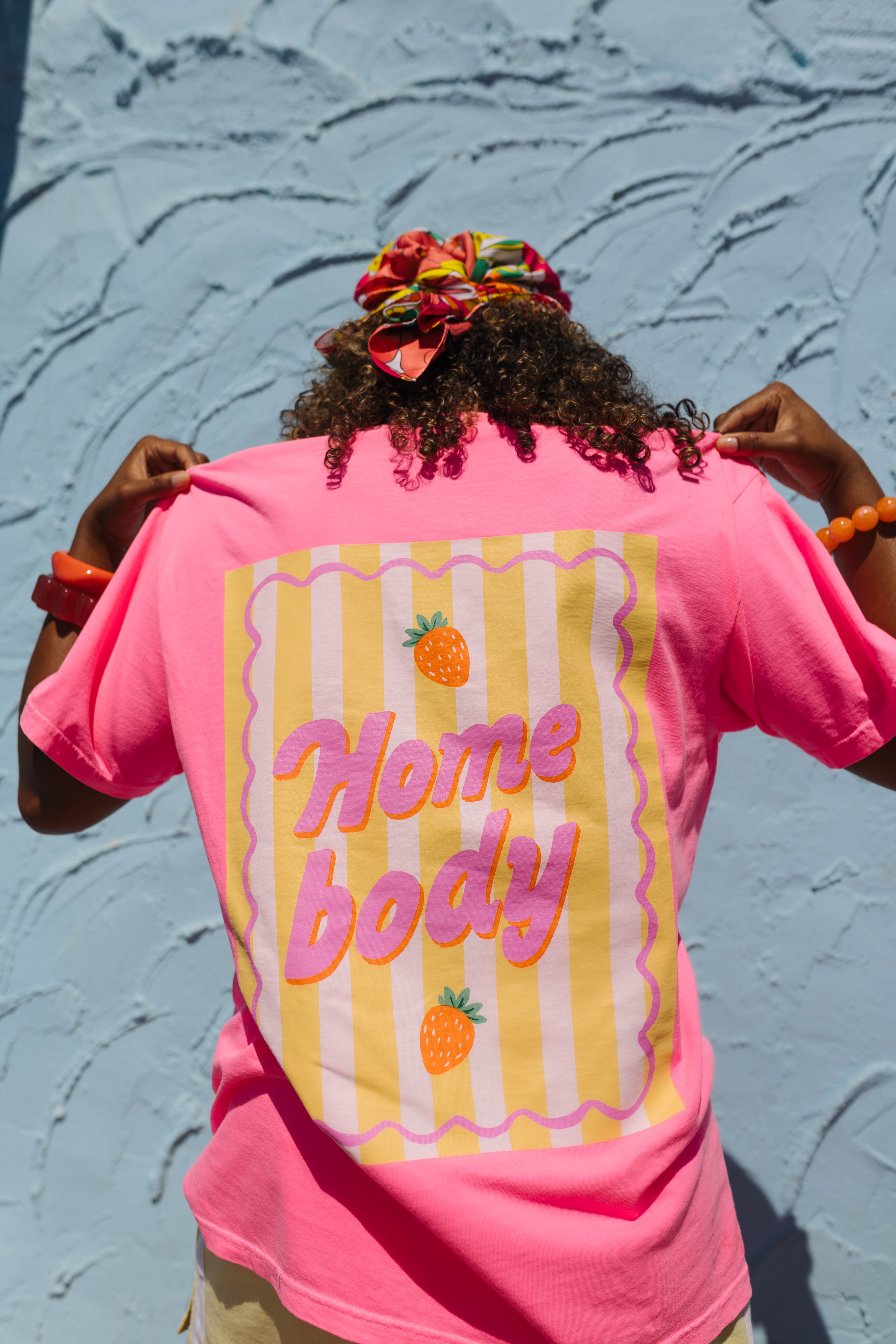 A person viewed from behind wearing a bright pink t-shirt with a retro graphic on the back. The graphic features yellow and white vertical stripes, wavy pink borders, and the words 'Home body' in pink bubble letters, accented with small orange pineapple illustrations.