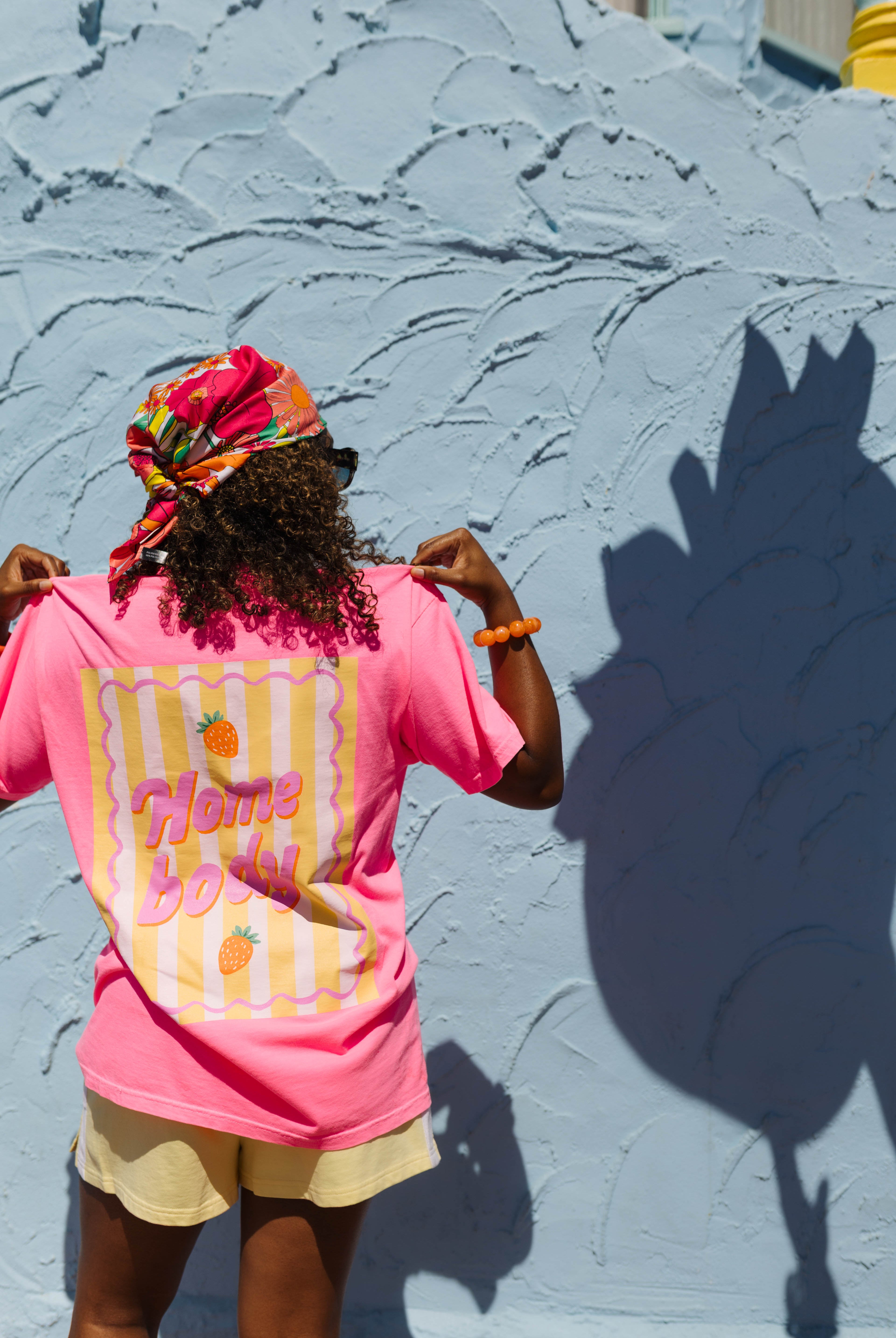 A person wearing a bright pink graphic t-shirt and a colorful headwrap stands with their back to the camera, posing in front of a textured light blue wall that casts a large shadow of the strawberry