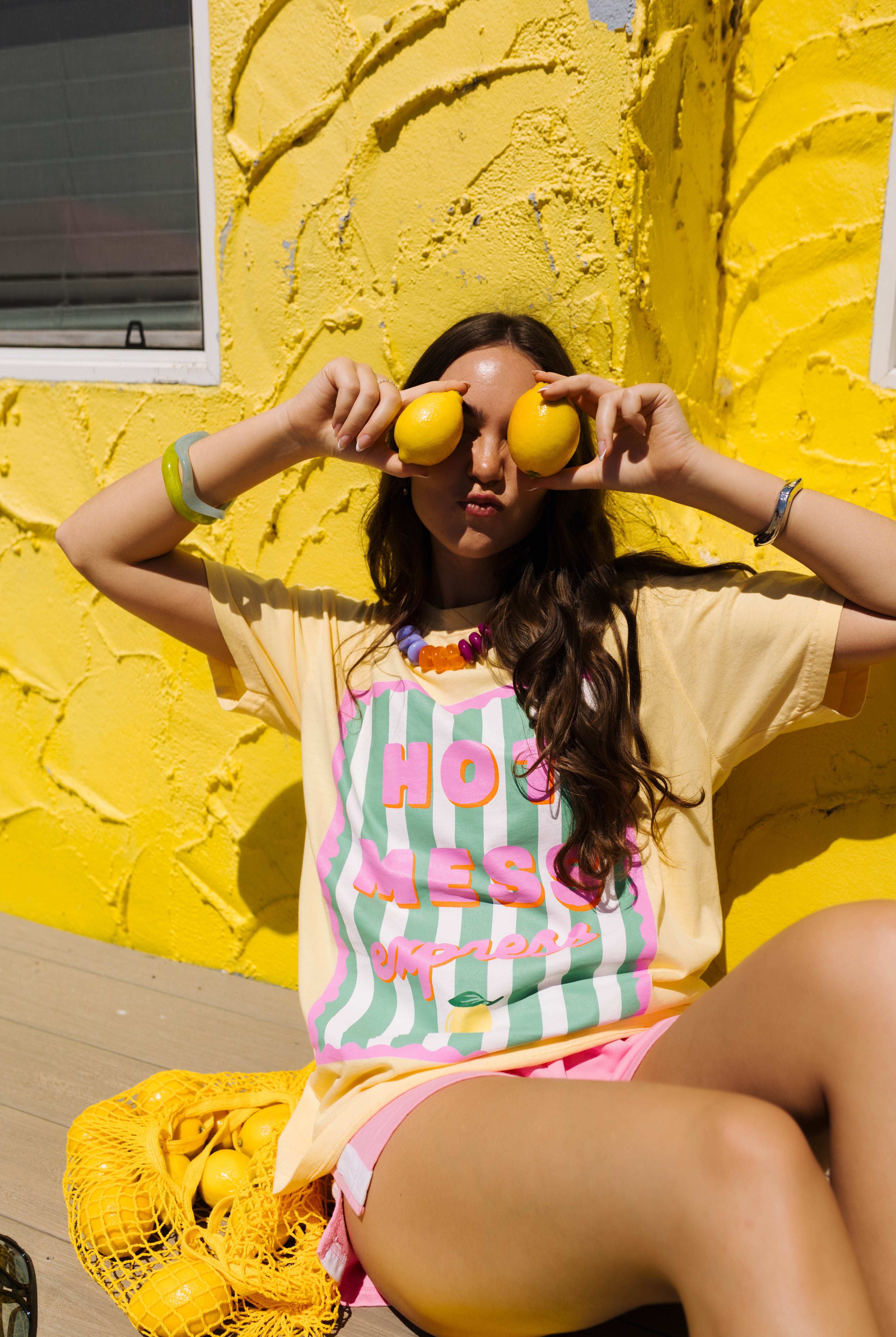 A woman sitting against a bright yellow stucco wall, wearing a pale yellow graphic T-shirt with pink and mint green stripes and the words 'HOT MESS' in pink script. She is holding two yellow lemons in front of her eyes, with a yellow mesh bag of lemons on the floor beside her.
