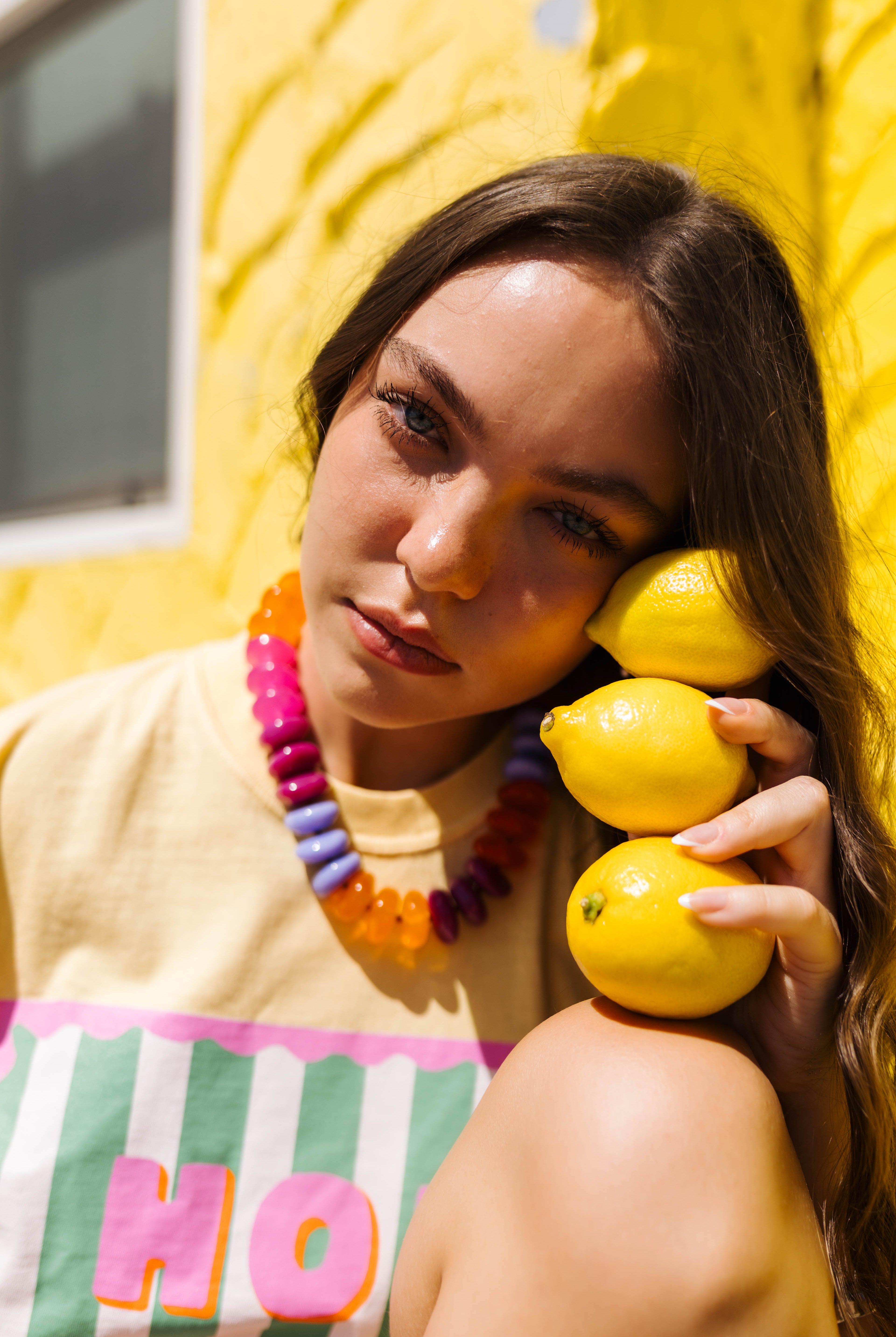 A woman with long brown hair leans against a bright yellow wall, holding three yellow lemons to her face. She is wearing a beige top with a colorful graphic print and a chunky beaded necklace.