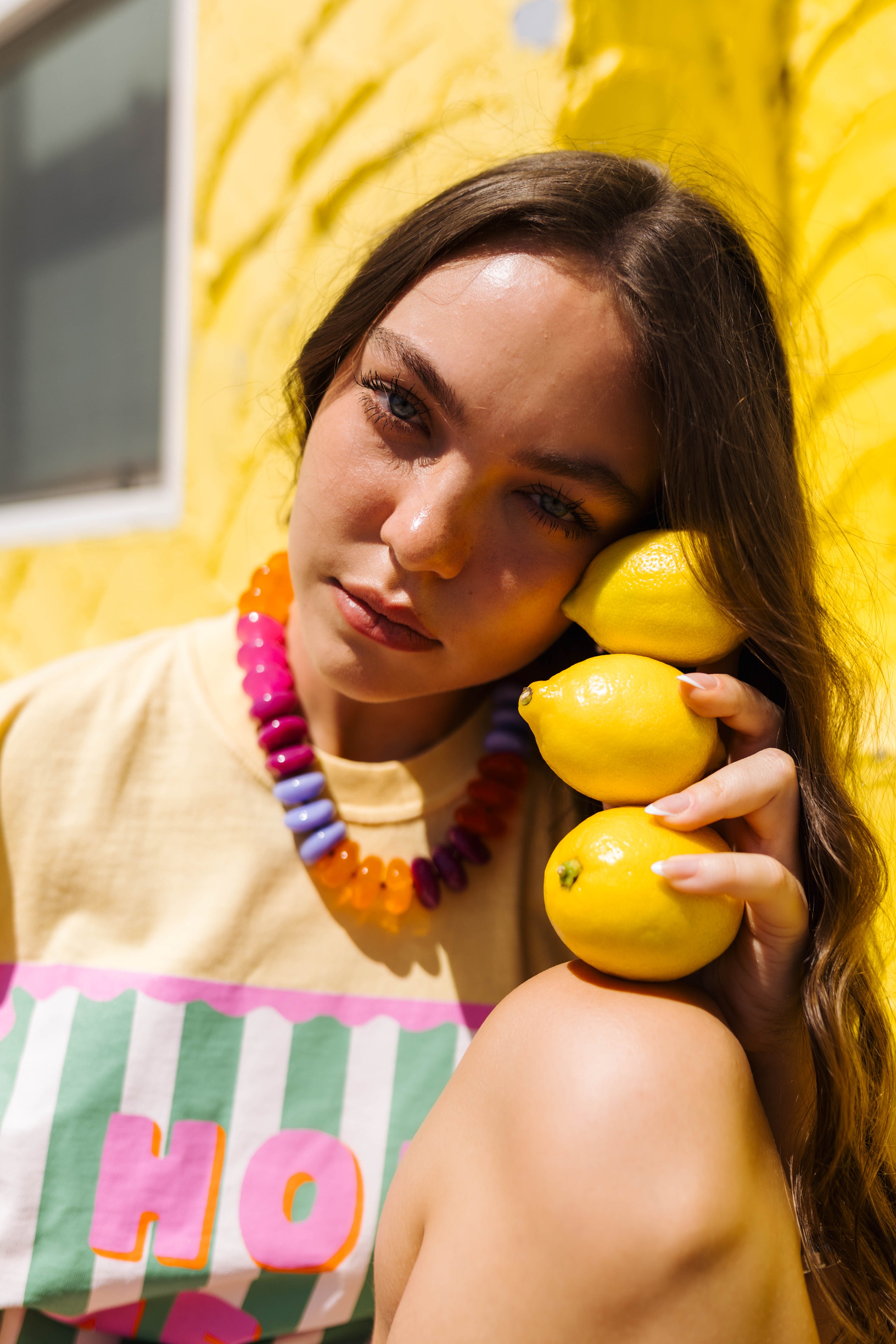 A woman with long brown hair leans against a bright yellow wall, holding three yellow lemons to her face. She is wearing a beige top with a colorful graphic print and a chunky beaded necklace.