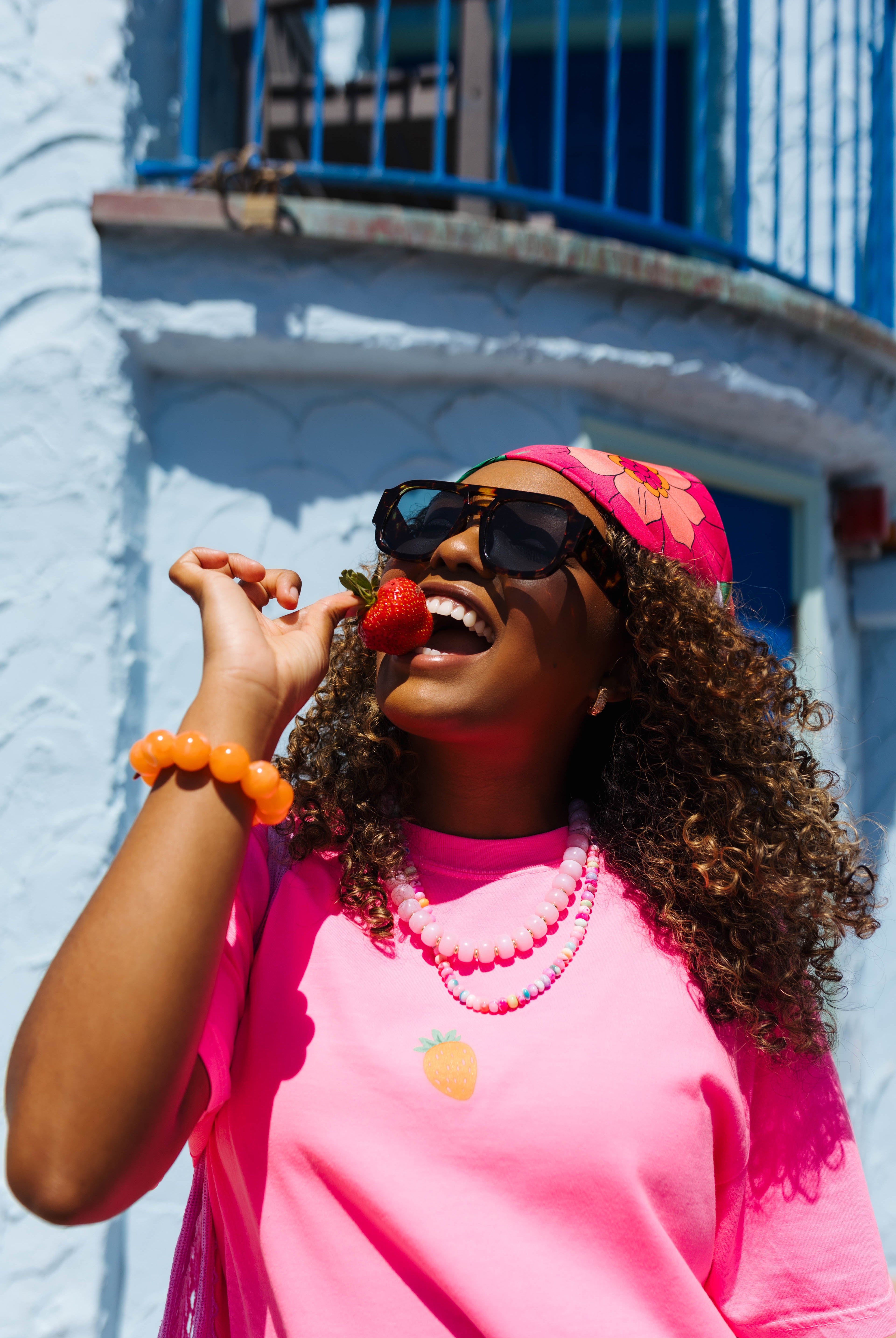 A woman wearing a bright pink t-shirt, matching pink headband, and sunglasses, holding a strawberry near her mouth. She is posed outdoors in front of a light blue stucco building with a blue metal balcony.