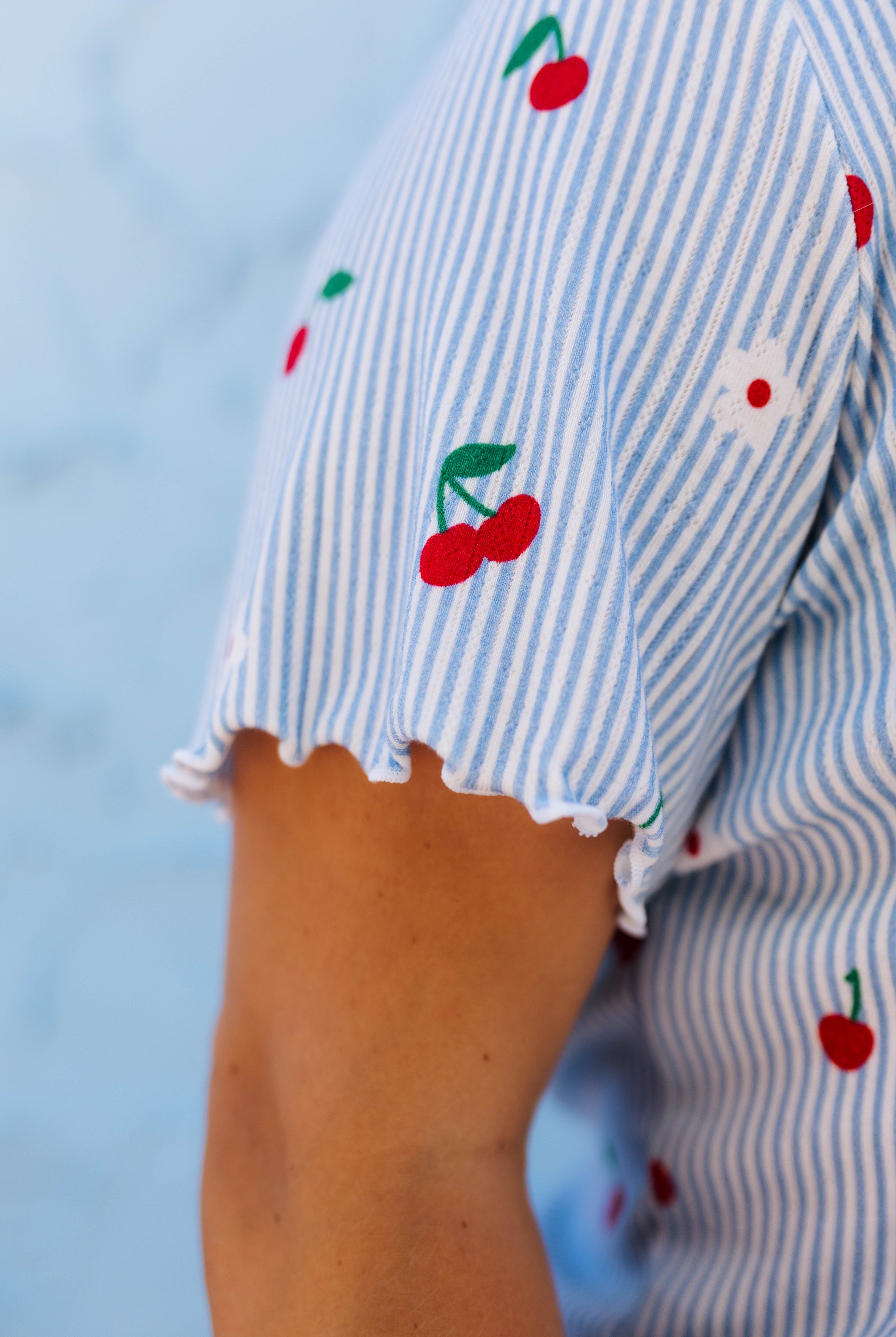 Close-up of a person wearing a light blue and white striped short-sleeve top with printed red cherries and a small white flower on the sleeve.