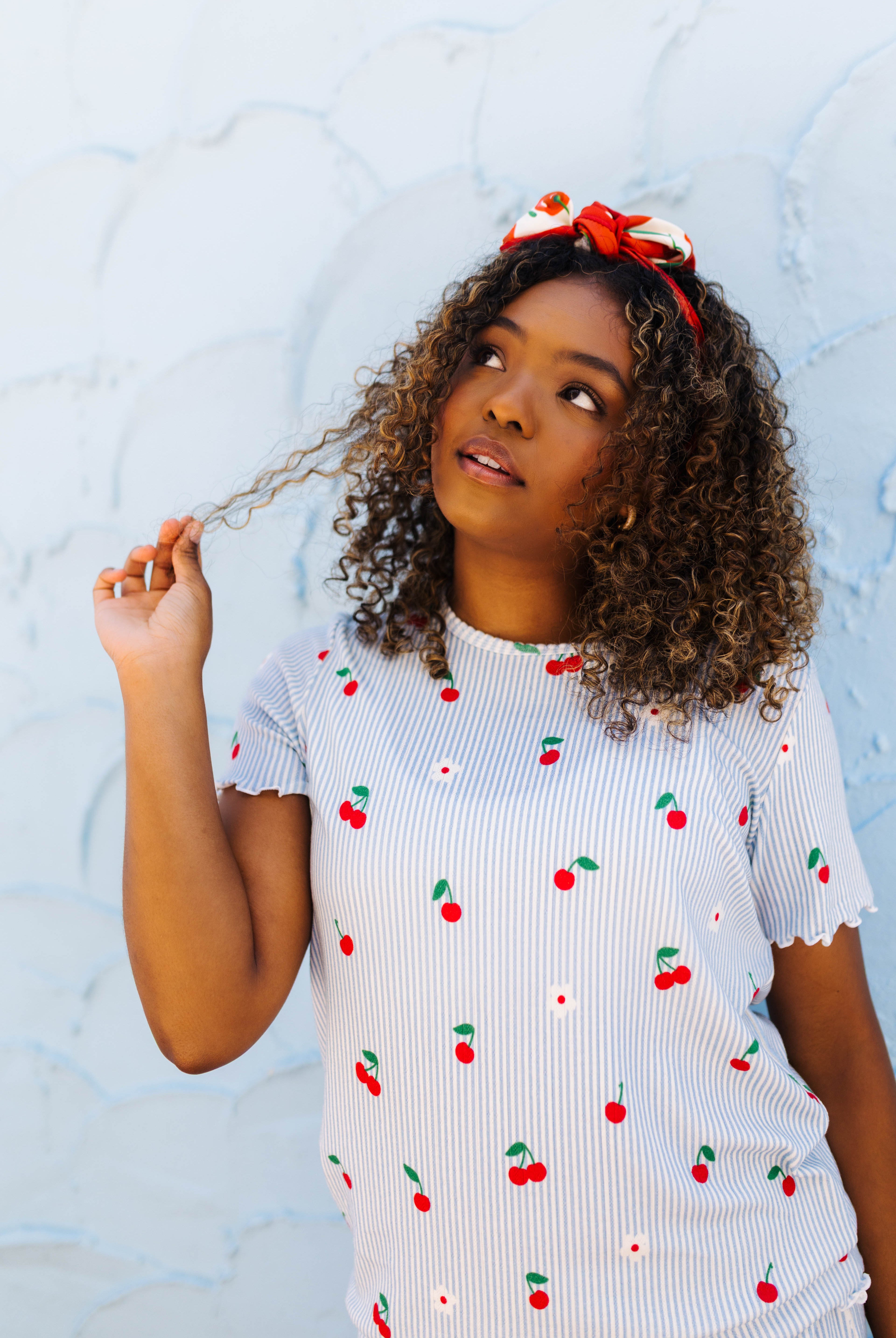 A woman wearing a white ribbed pj set with a red and green cherry print, paired with a red and white bow headband, posing against a textured light blue wall.