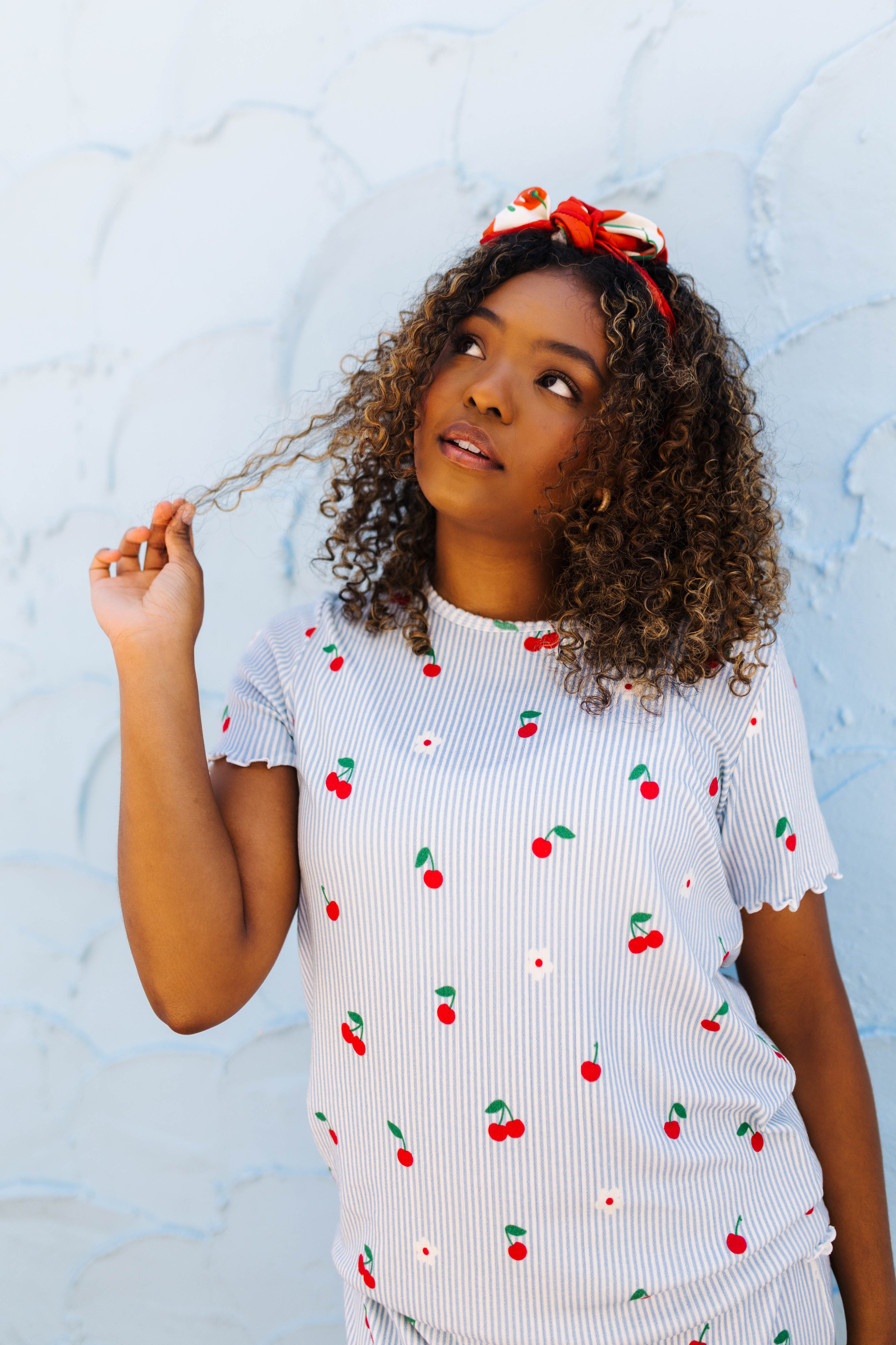 A woman wearing a white ribbed pj set with a red and green cherry print, paired with a red and white bow headband, posing against a textured light blue wall.