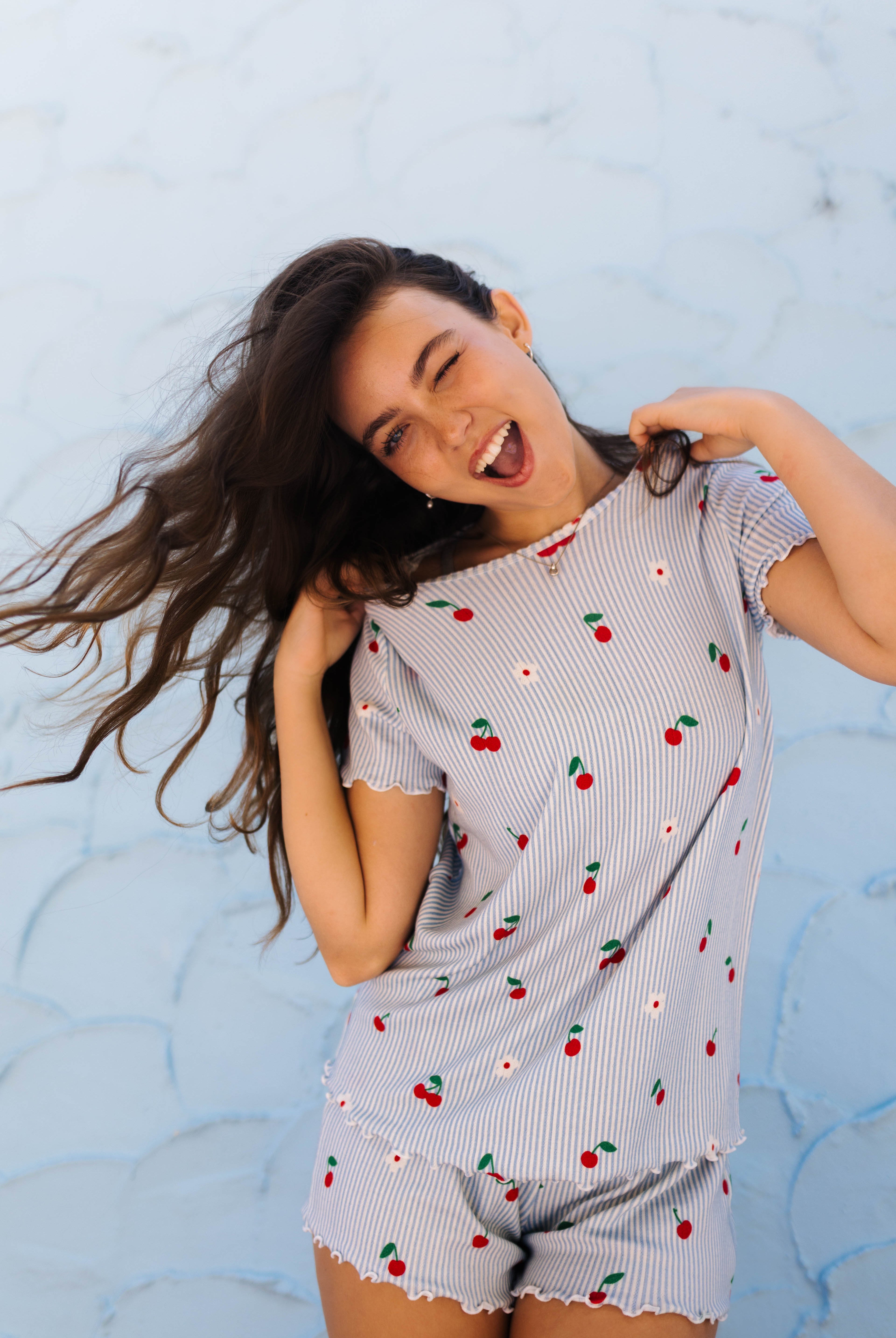A woman modeling a light blue and white striped short-sleeve pajama set with a cherry and daisy print, posing against a light blue textured wall.