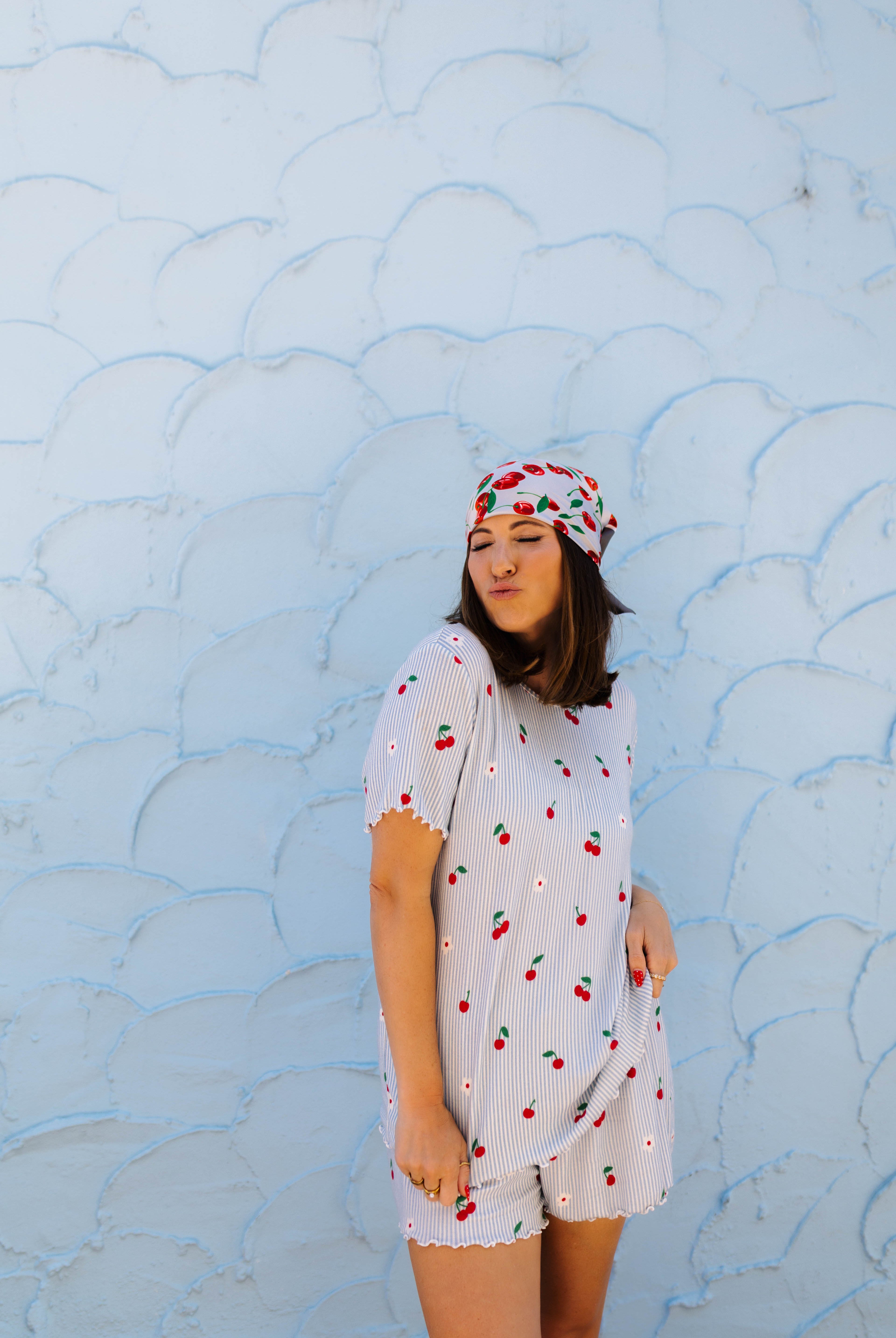 A woman wearing a white cherry-patterned short-sleeve pj and matching headscarf stands against a light blue textured wall.