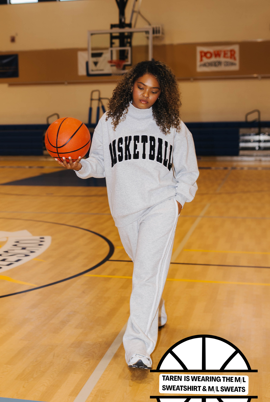 A woman stands on an indoor basketball court wearing a white sweatshirt and sweatpants set with black lettering that reads 'BASKETBALL'. She is holding an orange basketball.